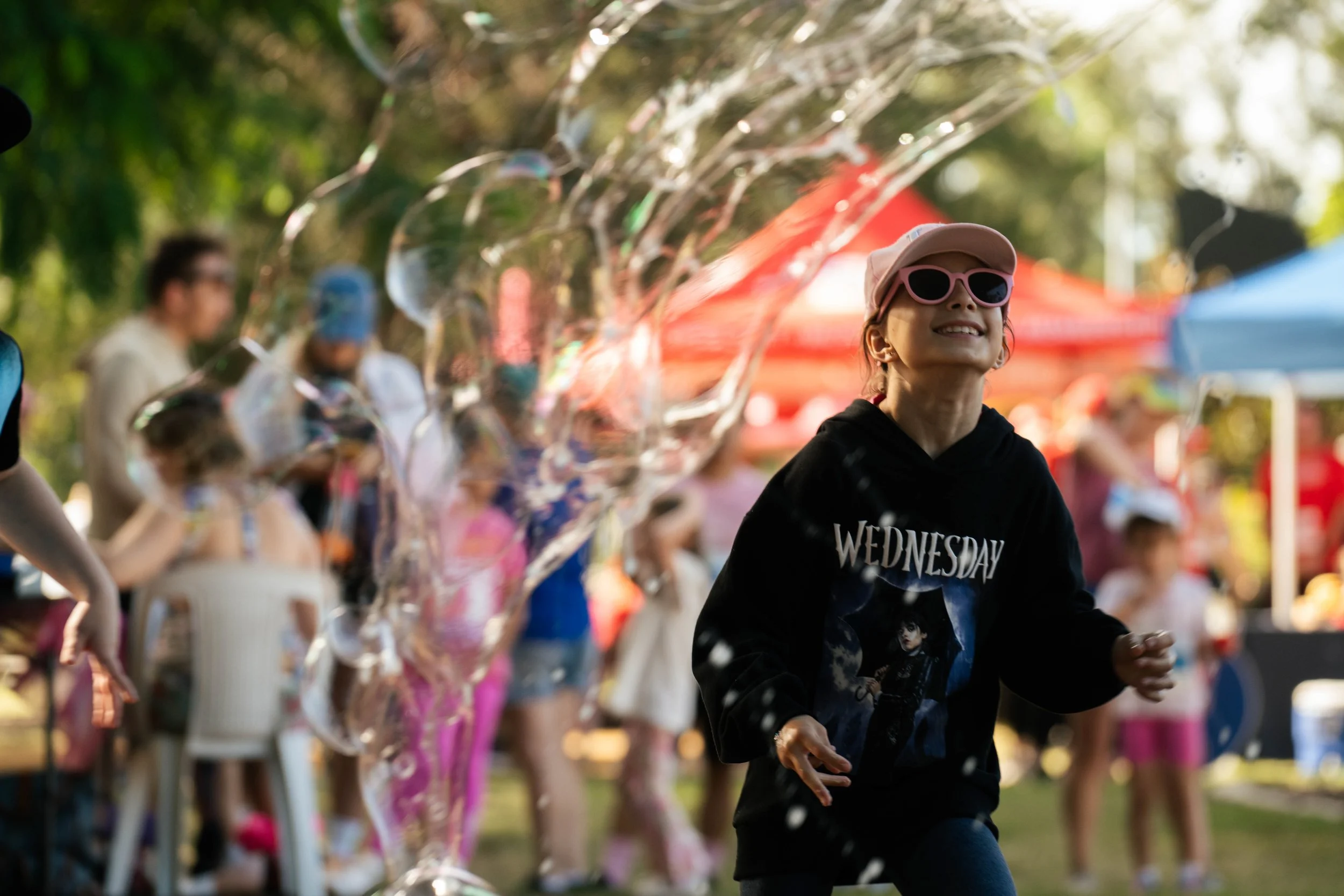 A young girl wearing pink sunglasses, a beige cap, and a black hoodie with a graphic print and the word 'Wednesday' on it, smiling and running outdoors during a daytime event with other people and tents in the background.