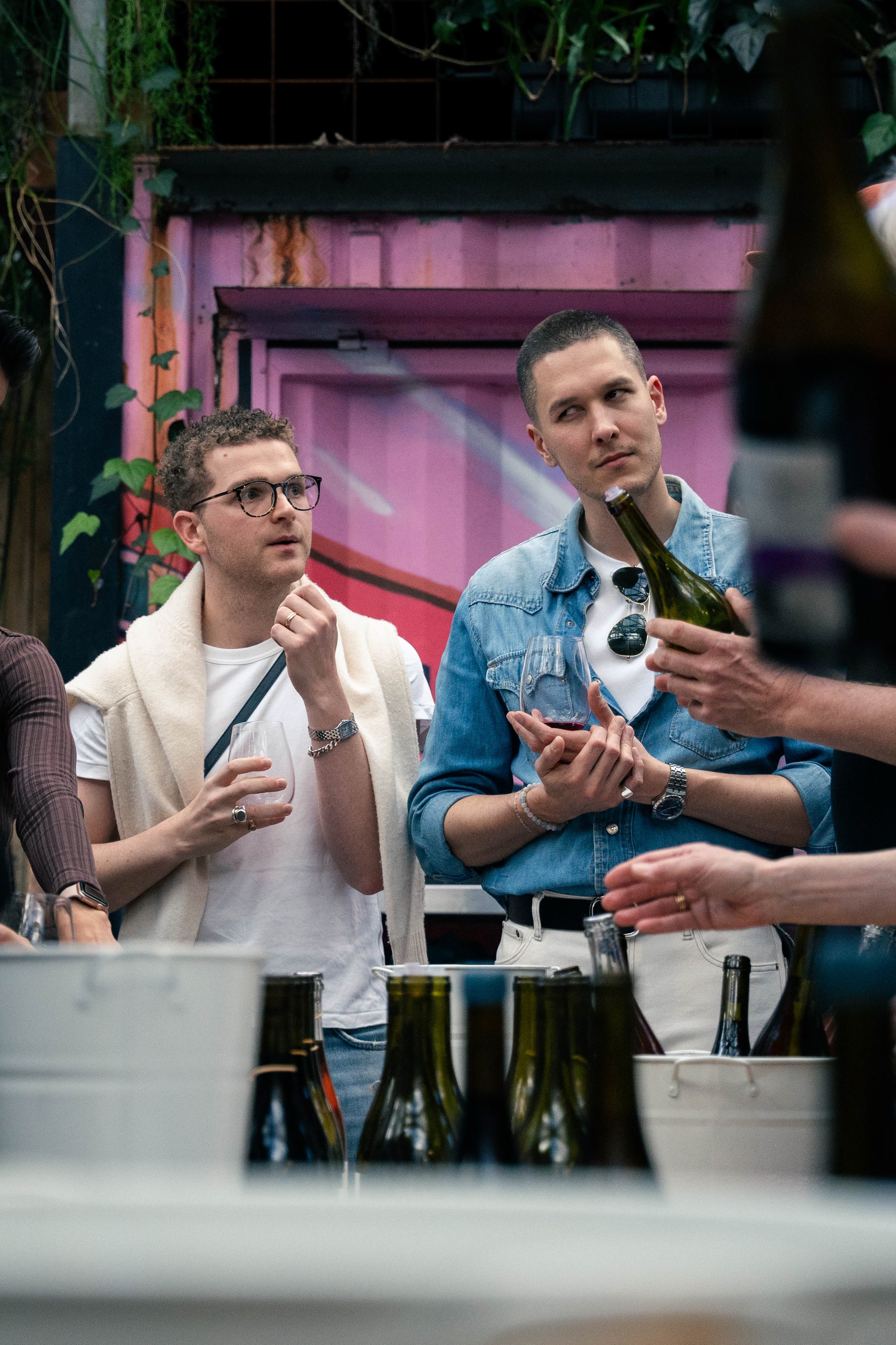 Two men at a social gathering holding wine glasses, one wearing glasses and a white shirt with a sweater draped over his shoulders, the other in a denim shirt, with bottles on the table in front of them.
