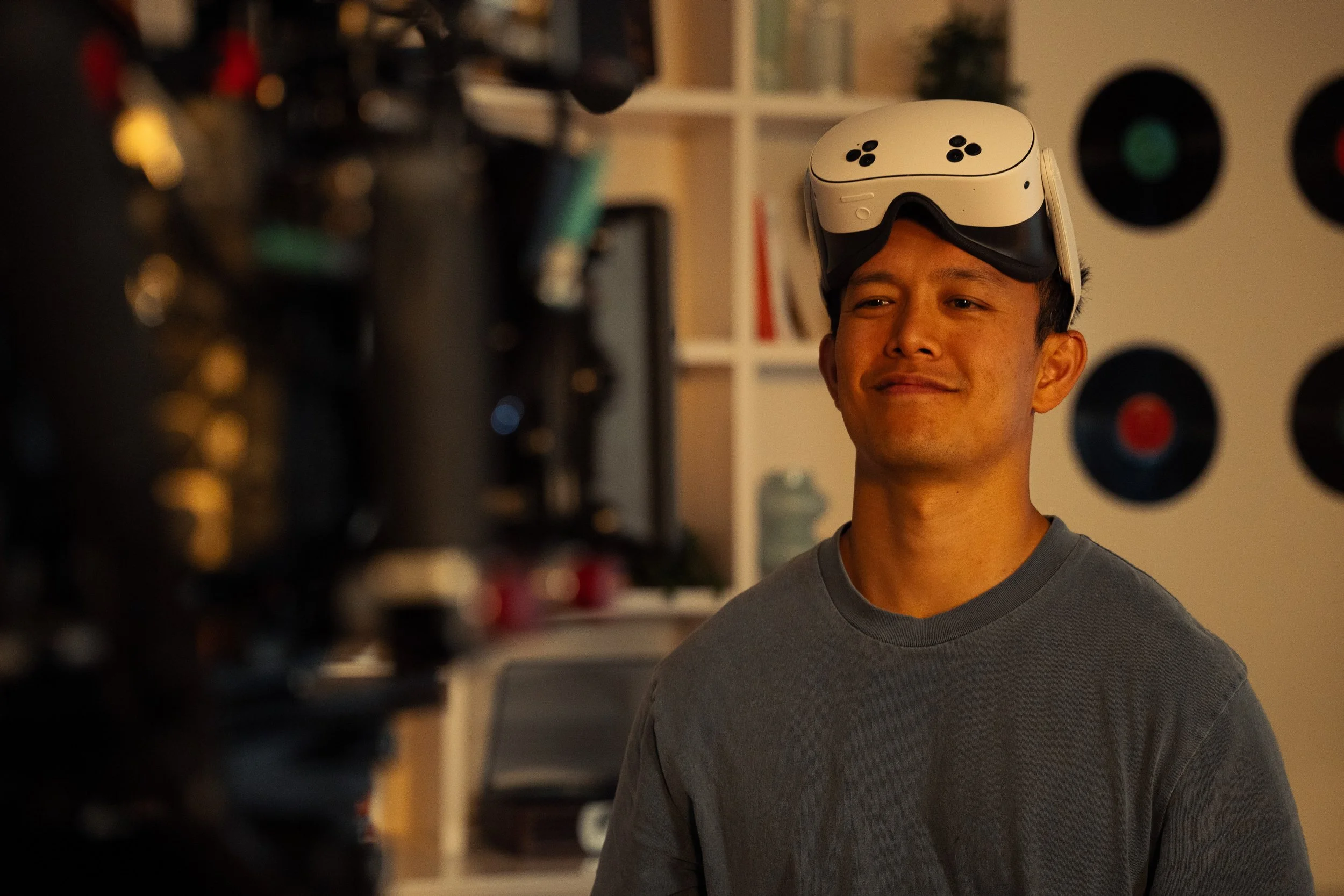 A young man with short hair wearing a grey T-shirt, balancing a virtual reality headset or goggles on his head, looking slightly to the side with a slight smile. The background shows a bookshelf and wall decor with vinyl records.