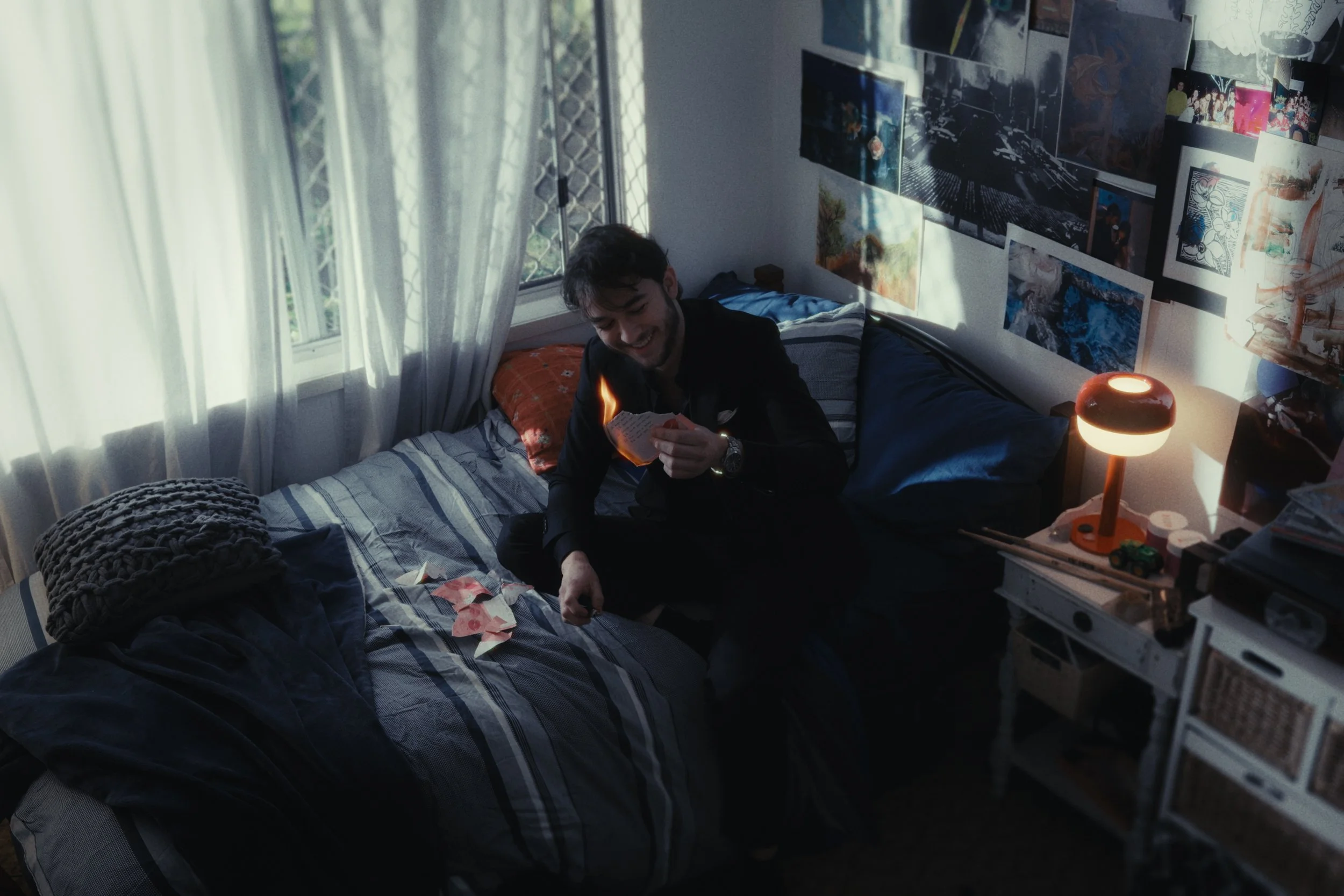A young man with dark hair and a beard sitting on a bed in a room, smiling and looking at a lit matchstick or small flame. The room has sunlight coming through a window with white curtains, and the walls are decorated with various pictures or posters