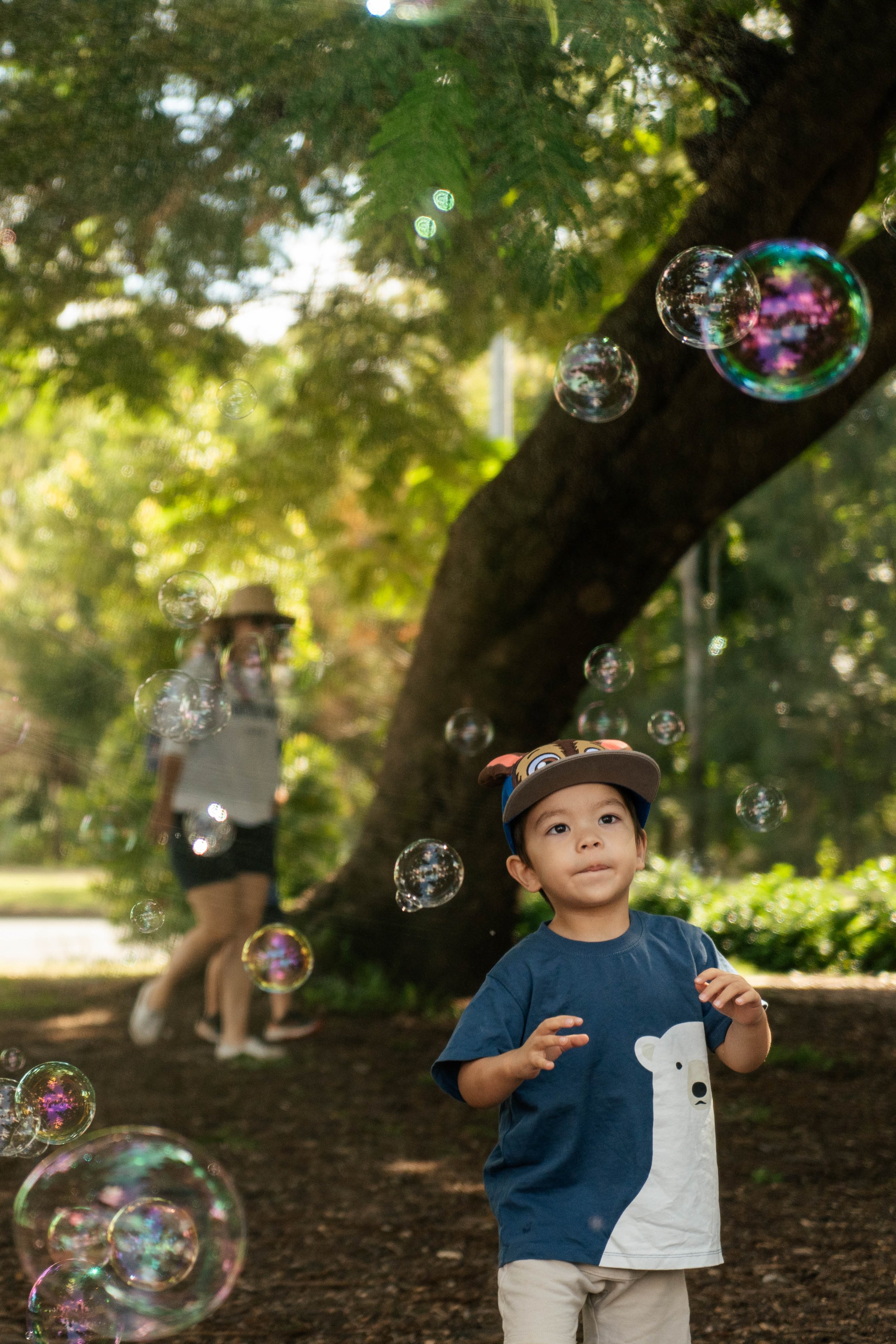 A young boy wearing a blue T-shirt with a bear graphic and a cartoon animal hat, standing outside among trees, with bubbles floating around him. In the background, a woman is walking, blurred.