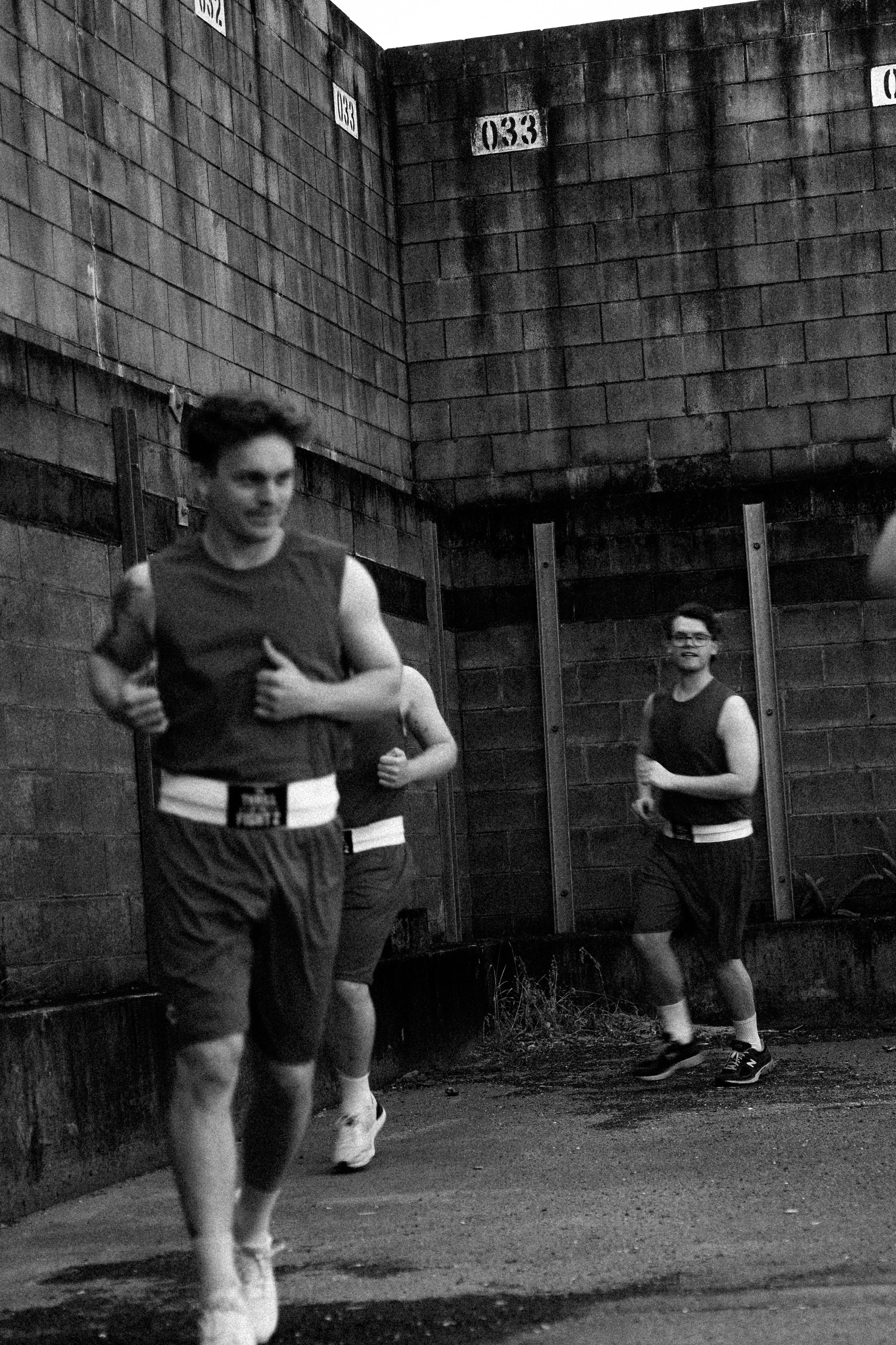 Black and white photo of three young men running outdoors along a wall with numbered markings, dressed in athletic clothing.