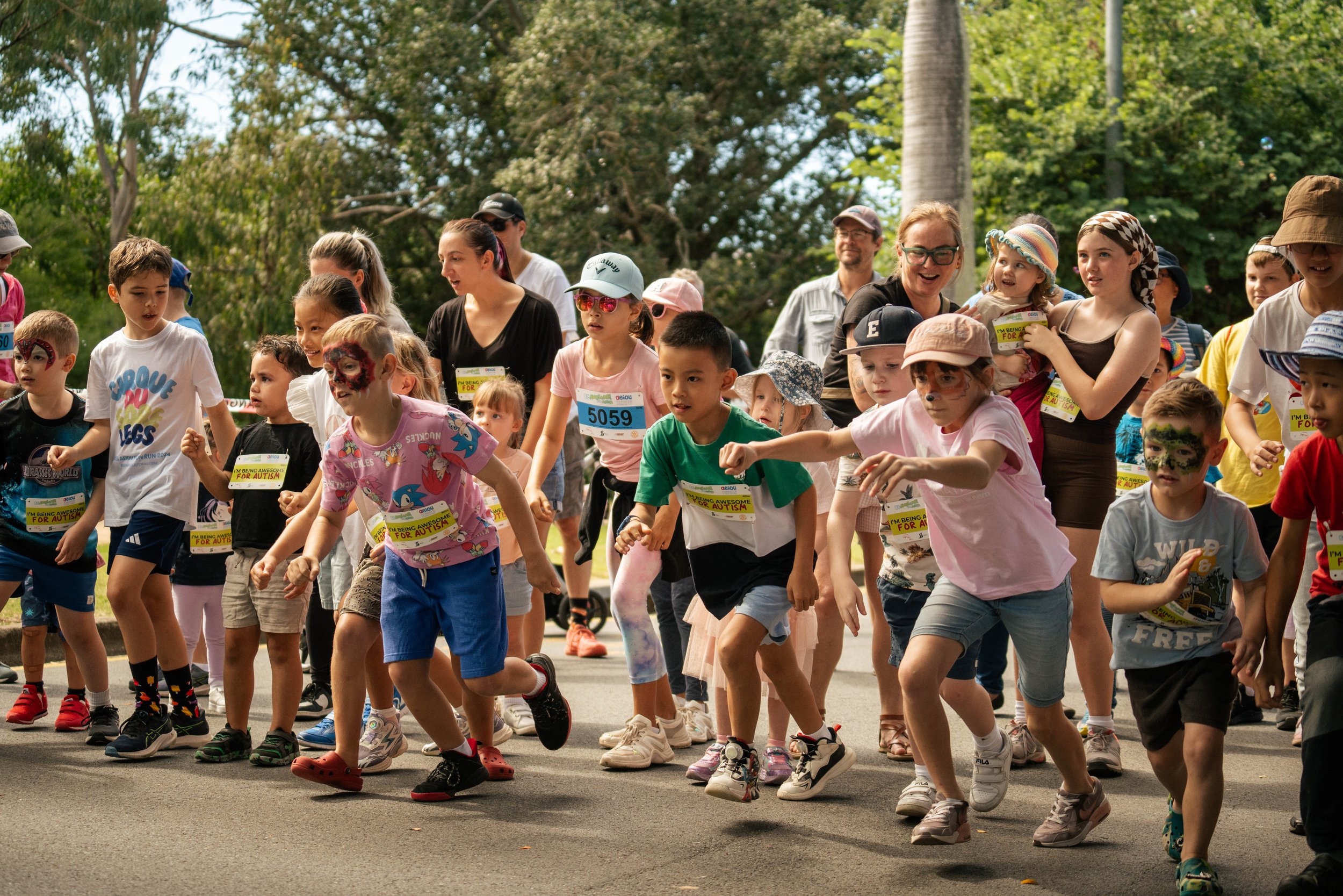 Children and adults participating in a fun run or race outdoors on a sunny day, with trees in the background.