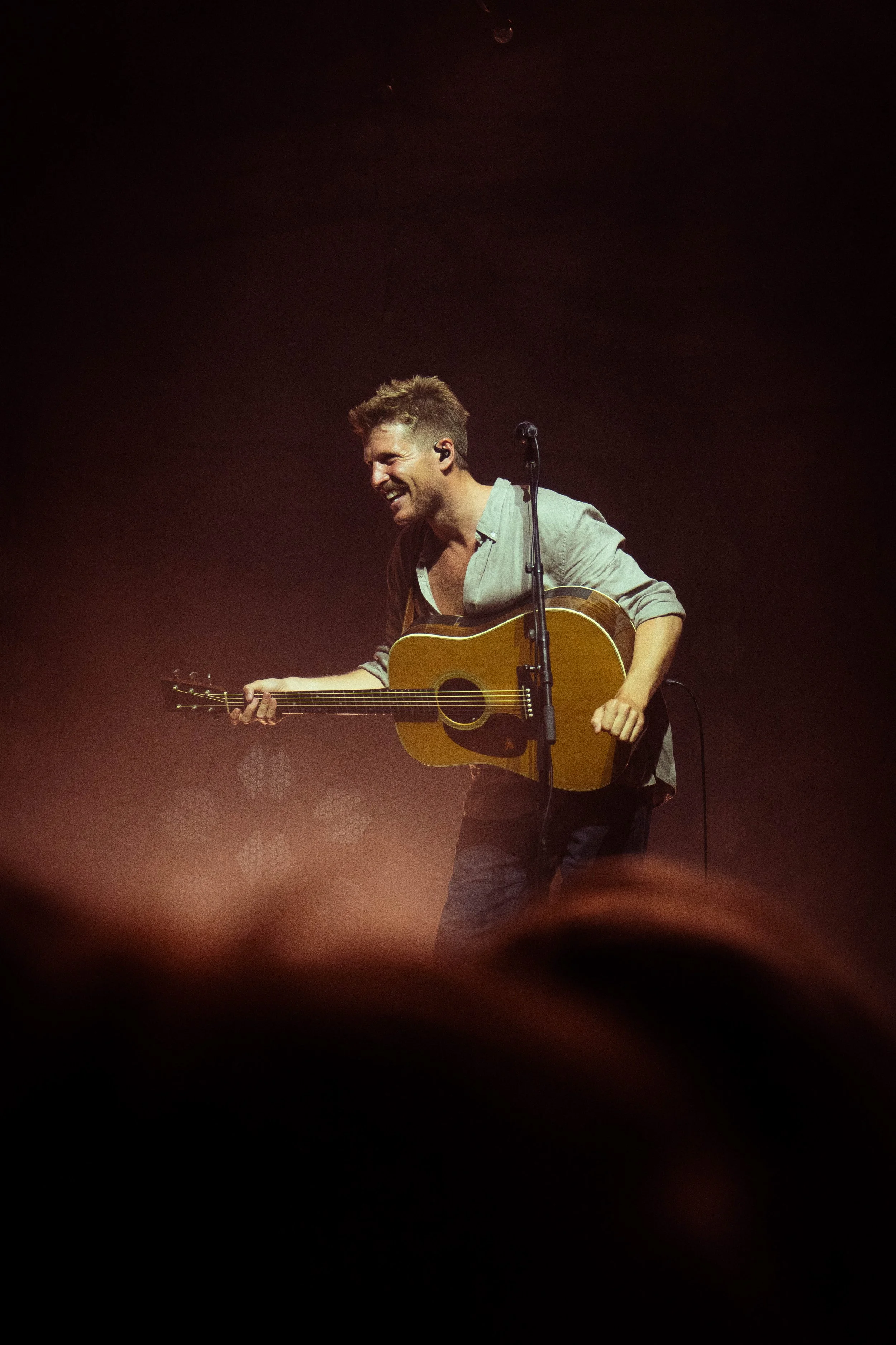 Man on stage playing an acoustic guitar, smiling, with dark background and stage lights.