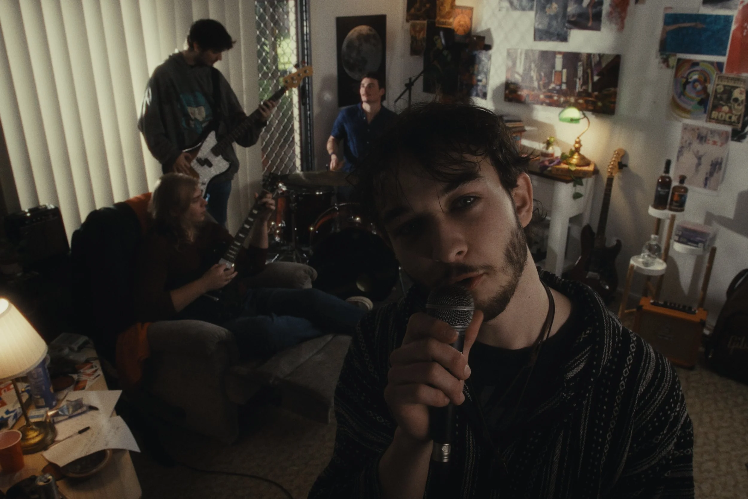Young man with dark hair and beard singing into a microphone in a room with four other people playing musical instruments, with posters and paintings on the walls.