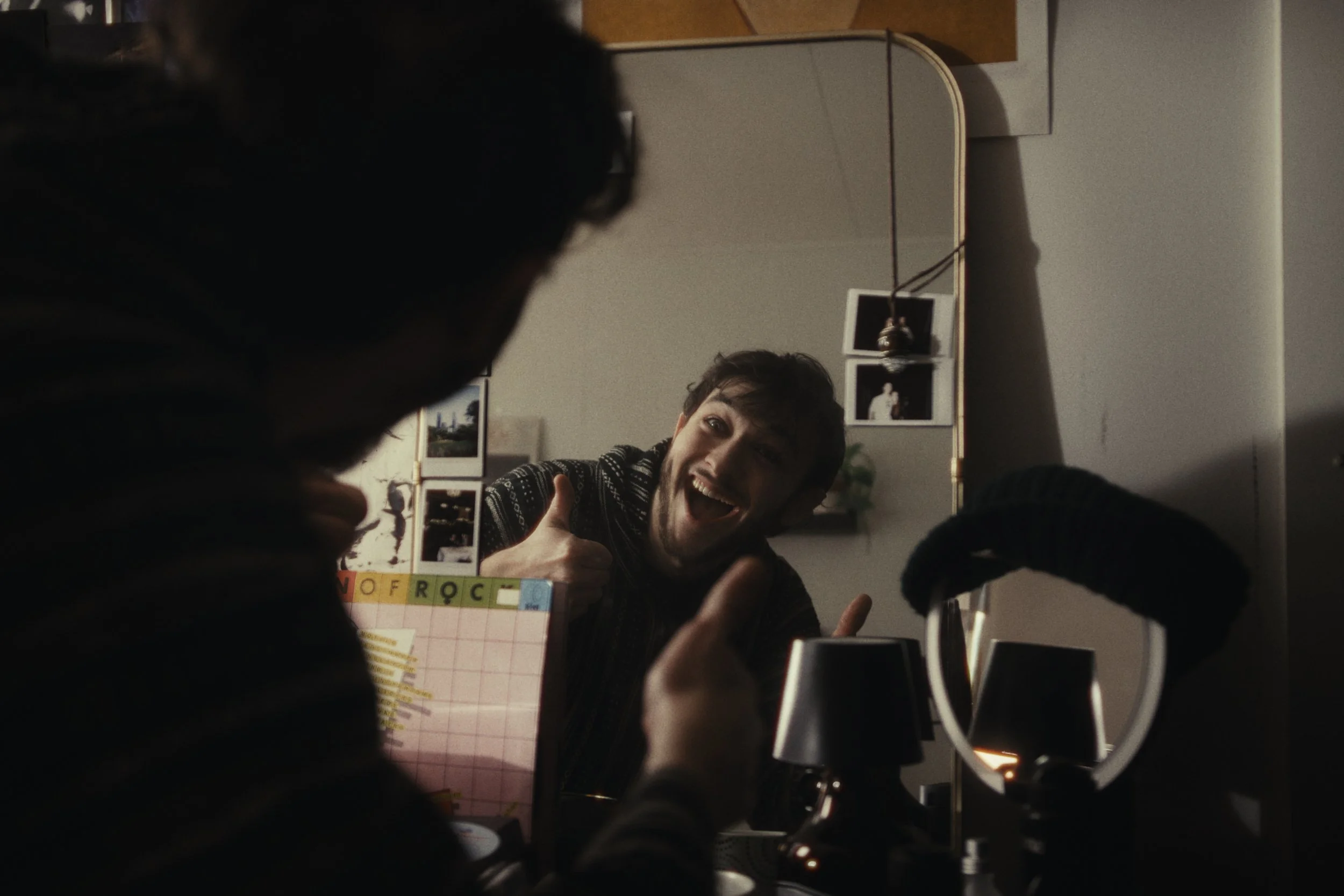 A young man with short hair giving a thumbs-up in front of a mirror, smiling. The reflection shows him sitting at a desk with various items, including a wall mirror, a lamp, and photographs on the wall behind him.