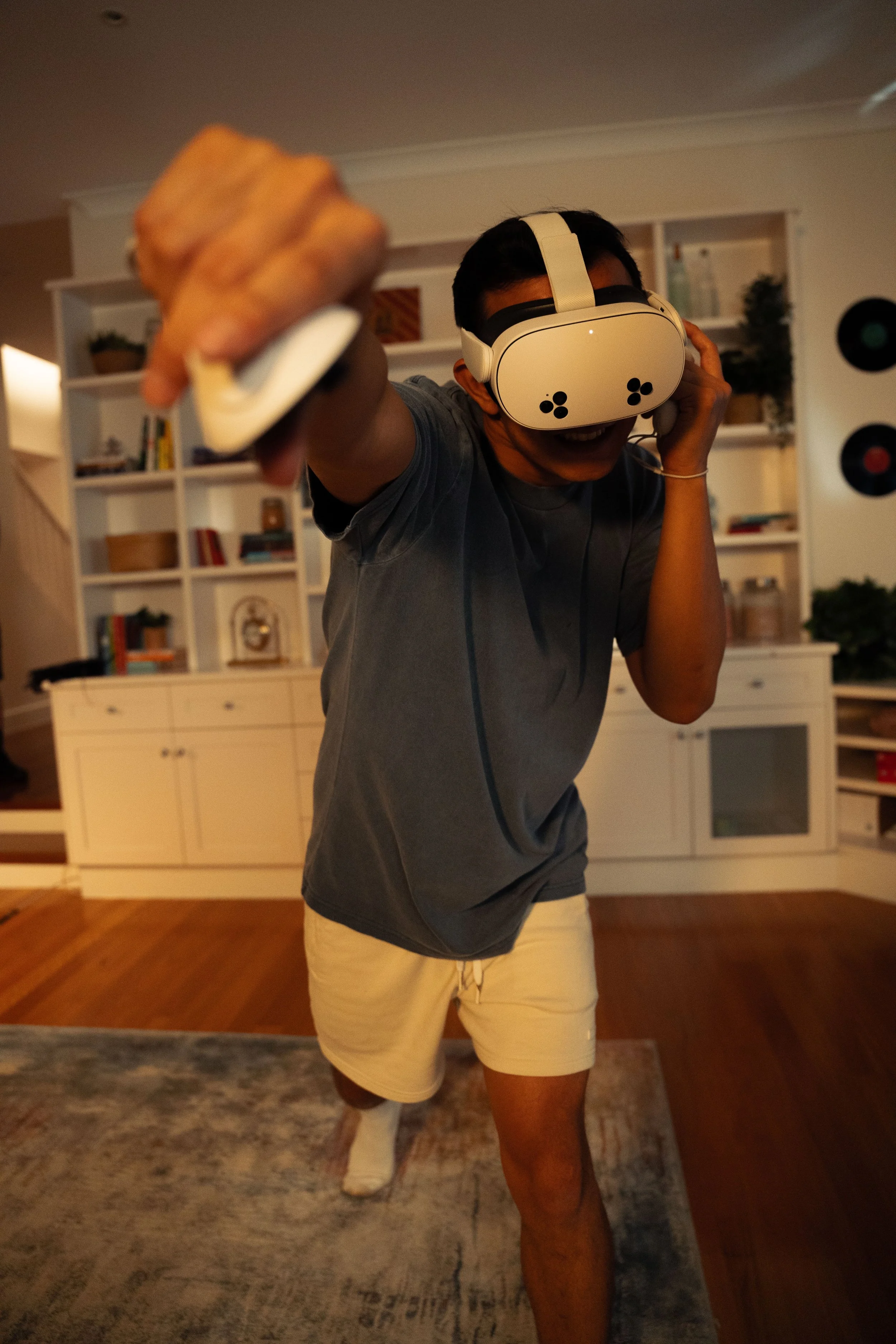 A young man wearing a virtual reality headset and headphones, standing on one leg with a fierce pose, in a living room with white shelving and wooden floors.