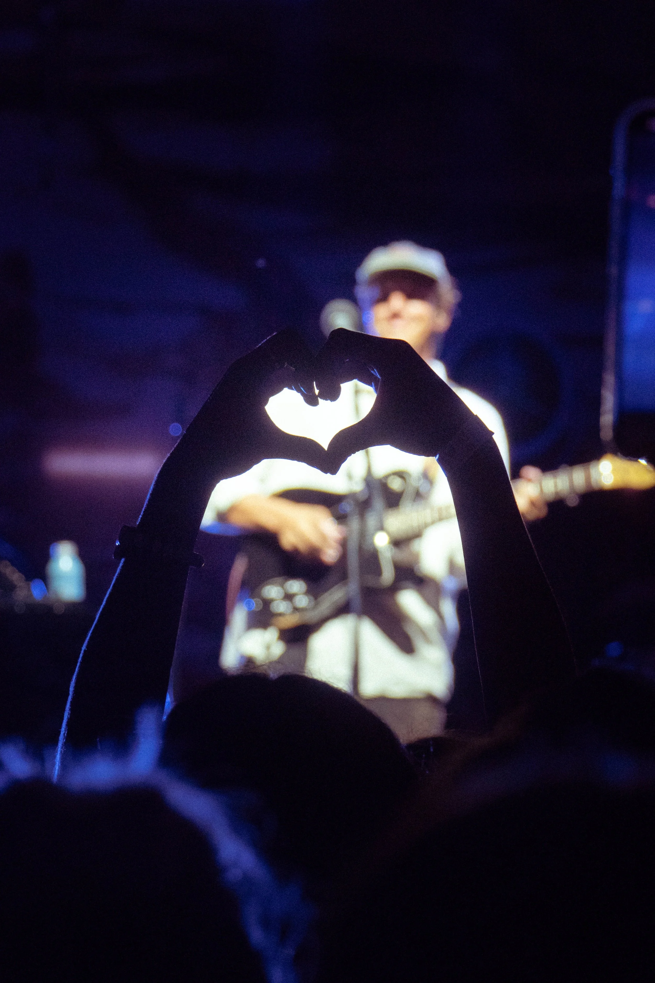 A concert scene with a silhouette of a person making a heart shape with their hands in the foreground and a musician playing guitar on stage in the background.