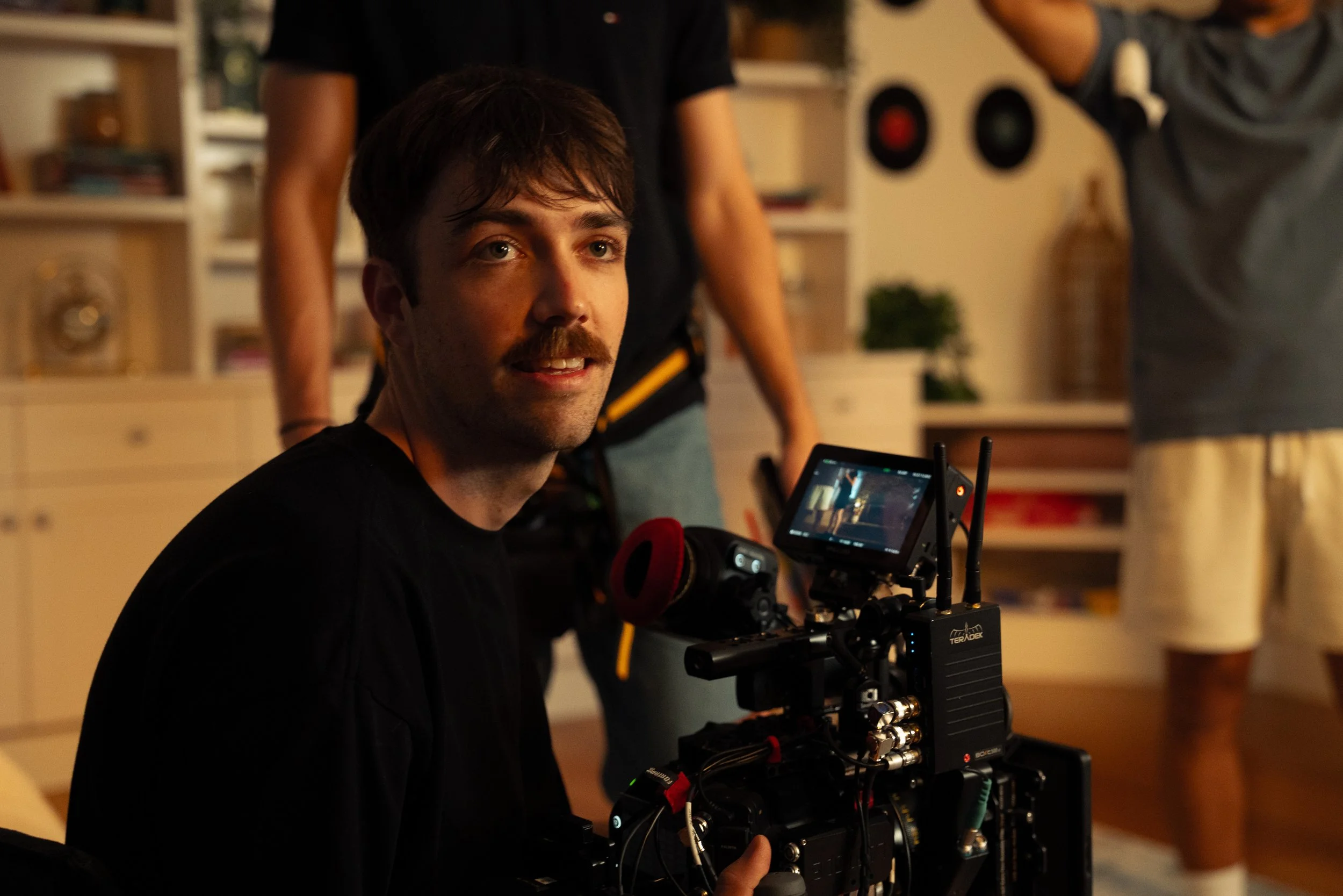 A man with a mustache, sitting on the floor, operating a professional video camera in an indoor setting. Two other people are in the background, one with a camera or phone taking a picture.