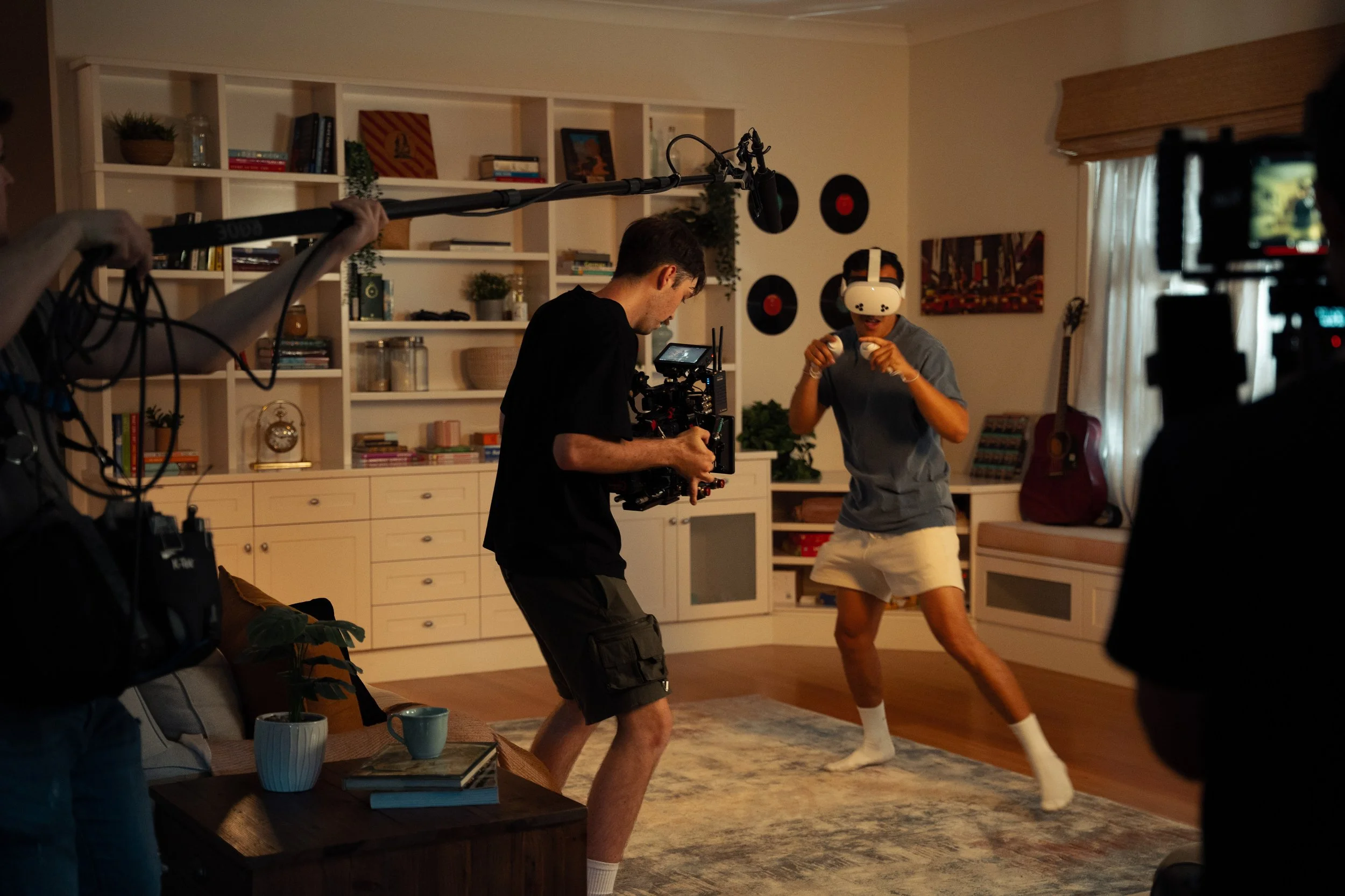 A person wearing a virtual reality headset is standing in a living room, posing in a boxing stance while being filmed by a camera crew with a camera operator and a person holding a boom microphone.