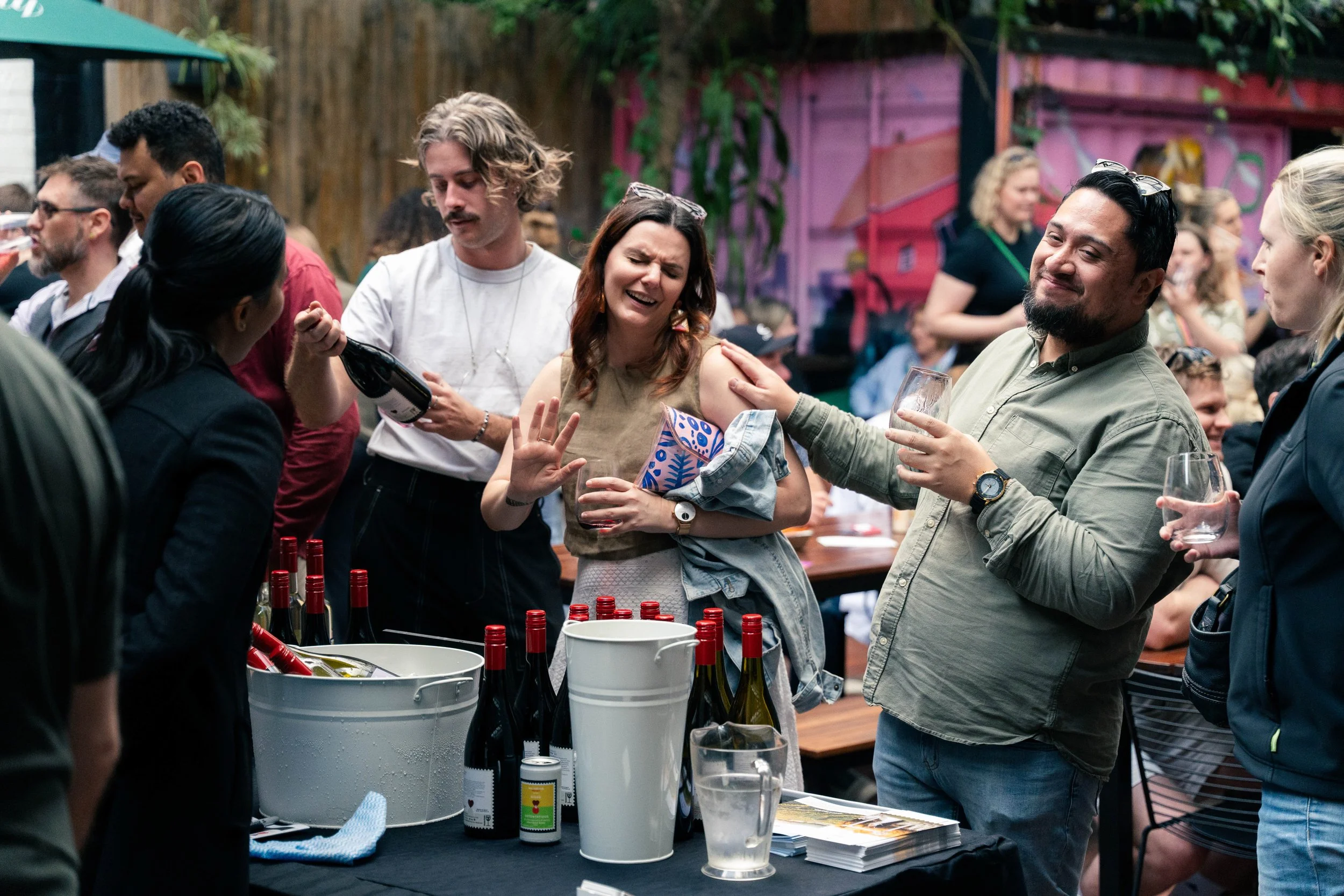 People at a social gathering or party, some of them holding drinks, with bottles of wine on a table in the foreground.