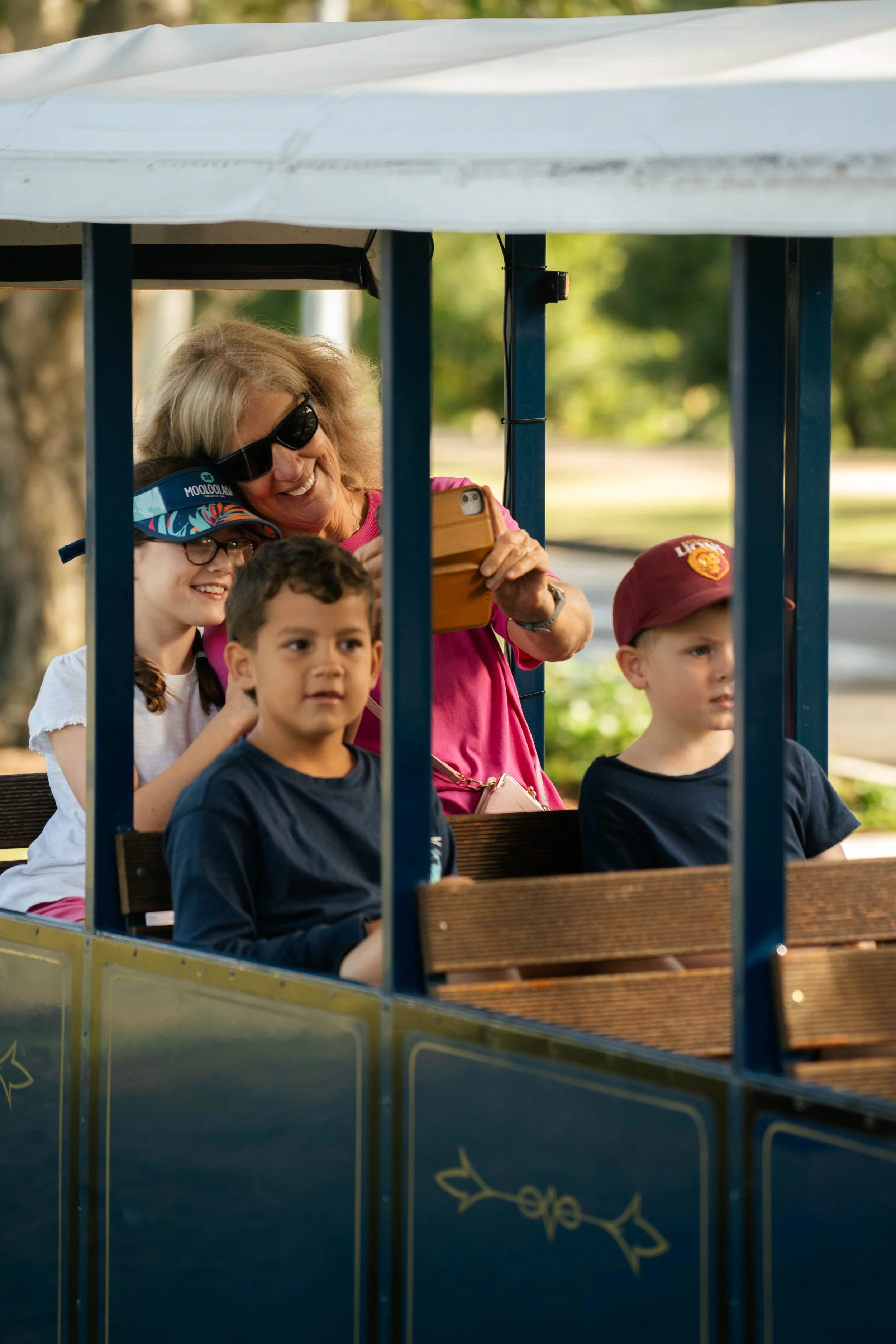 A woman and three children enjoying a ride on a small train at an outdoor amusement park or zoo, with green trees and path in the background.