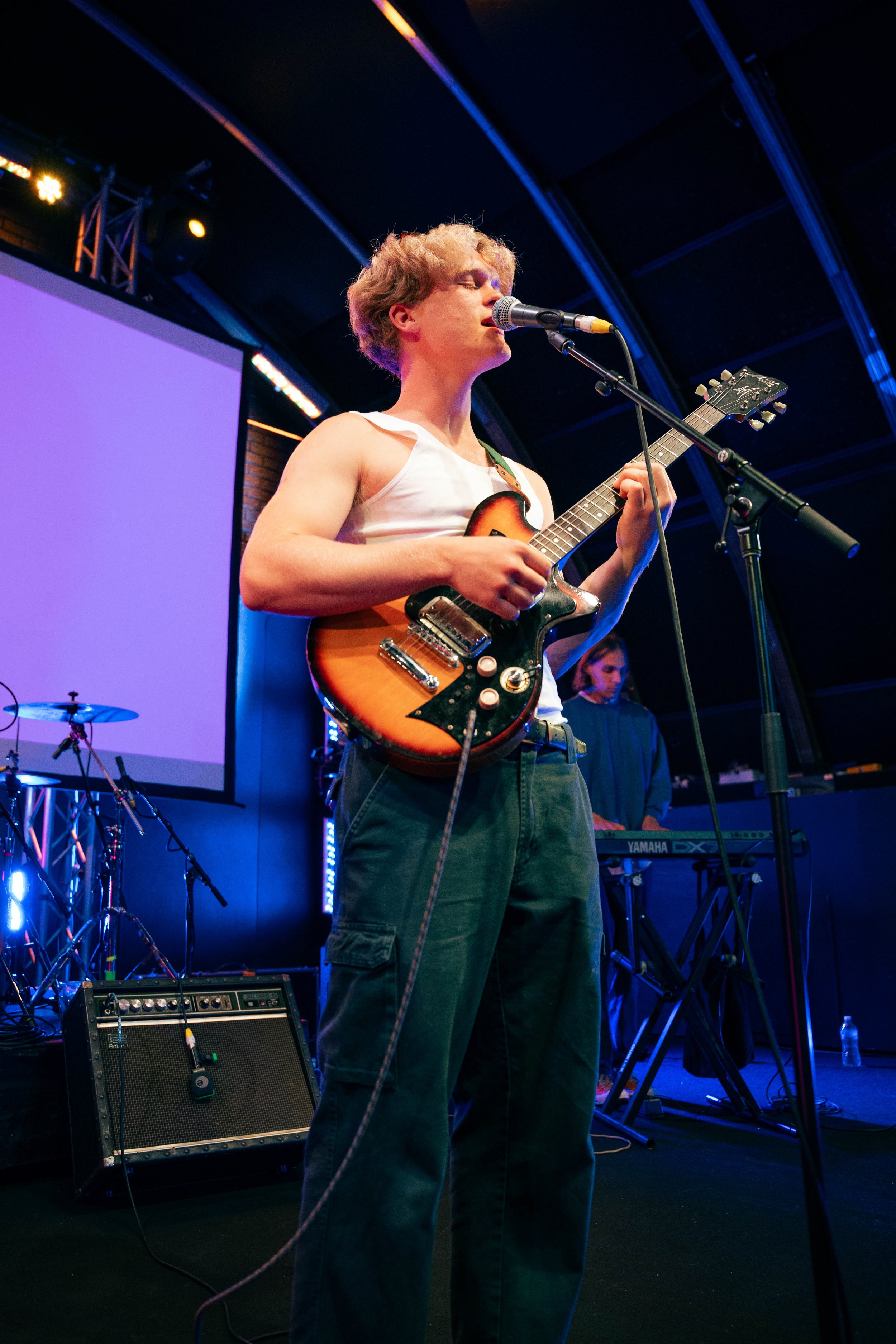 A young man with curly blonde hair performing on stage, singing into a microphone and playing an electric guitar, with a keyboard and drums in the background, under colorful stage lights.