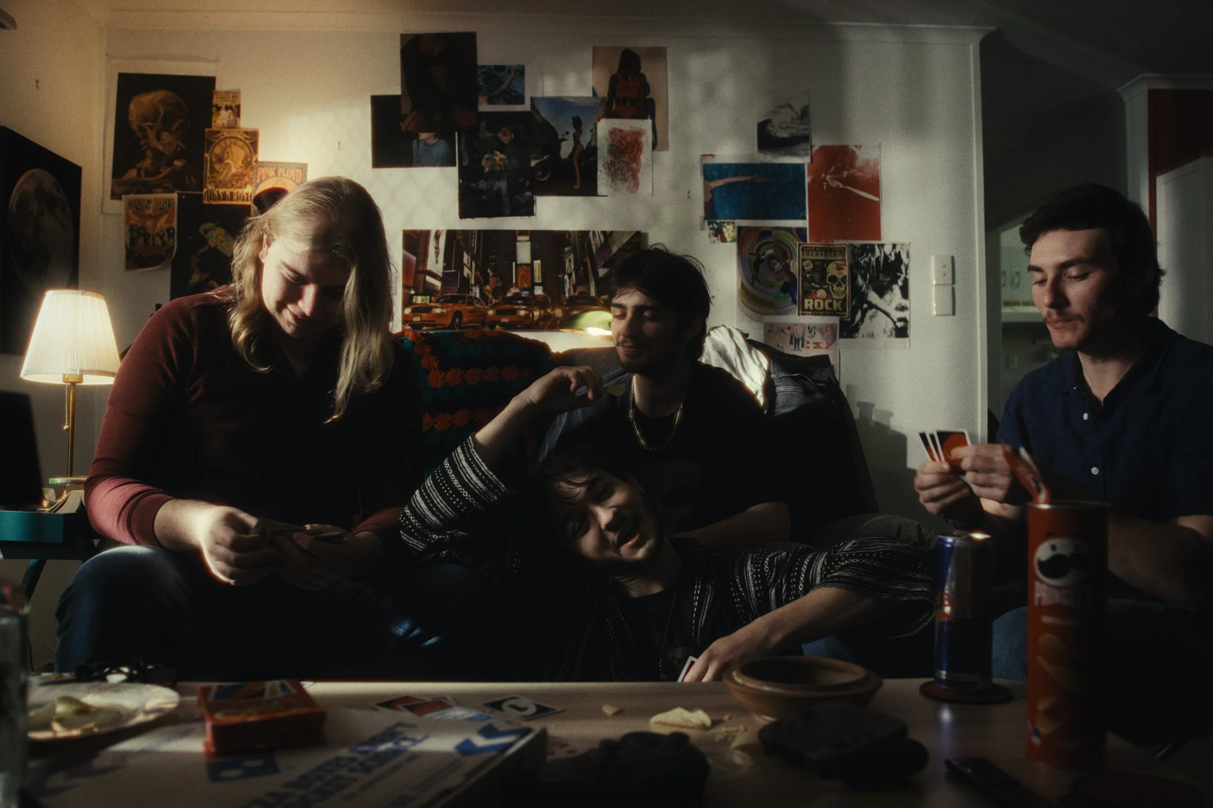 Four young adults are gathered in a dimly lit living room playing cards and having snacks, with a wall decorated with posters in the background.