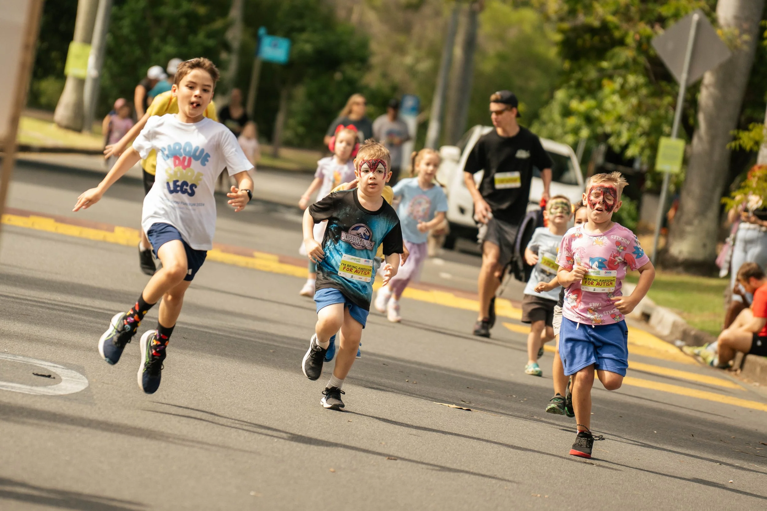 Children participating in a race, running on a street with some face paint and wearing casual clothes, with a man and other children in the background.
