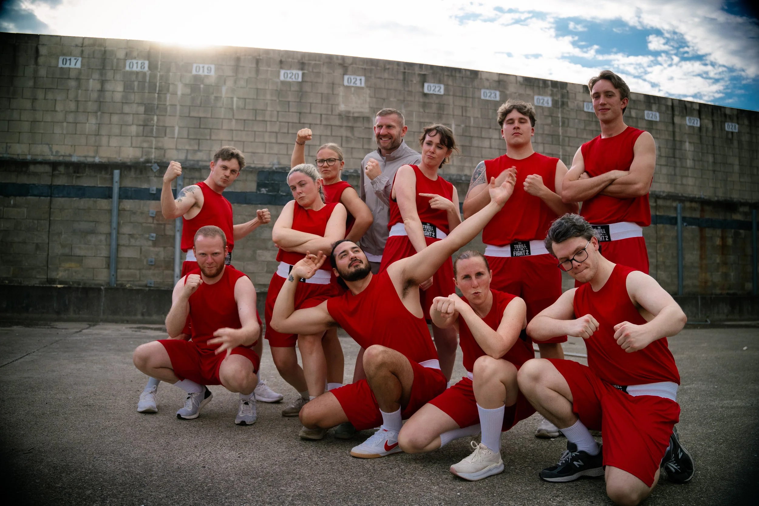A group of ten young adults in red athletic uniforms posing outdoors on a paved surface with a brick wall in the background. They are making various poses with confident expressions.