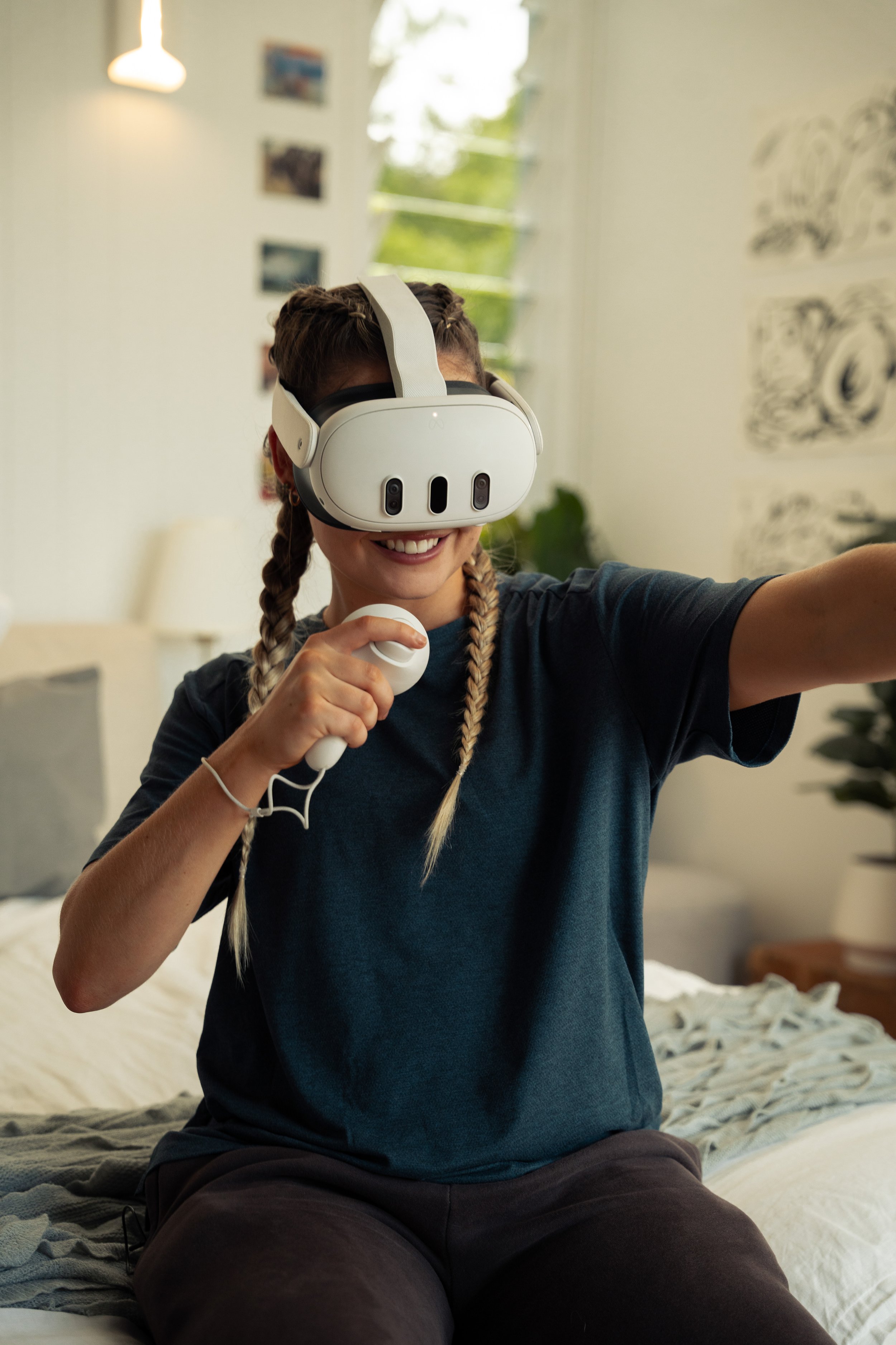 Woman wearing a virtual reality headset, smiling, sitting on a bed, holding a microphone, with a background of houseplant and wall decorations.