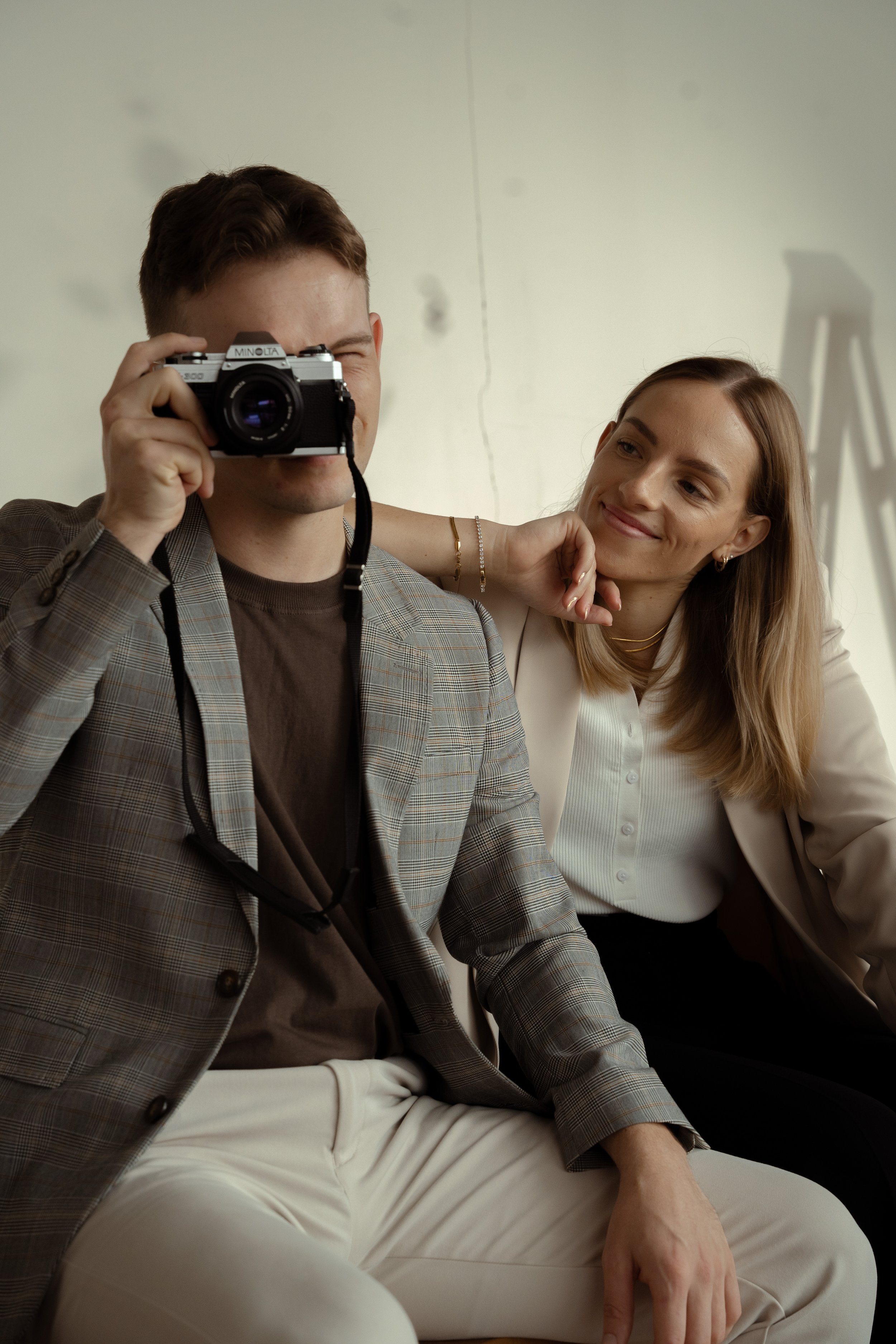 A man takes a selfie with a camera while a woman leans on his shoulder and smiles.