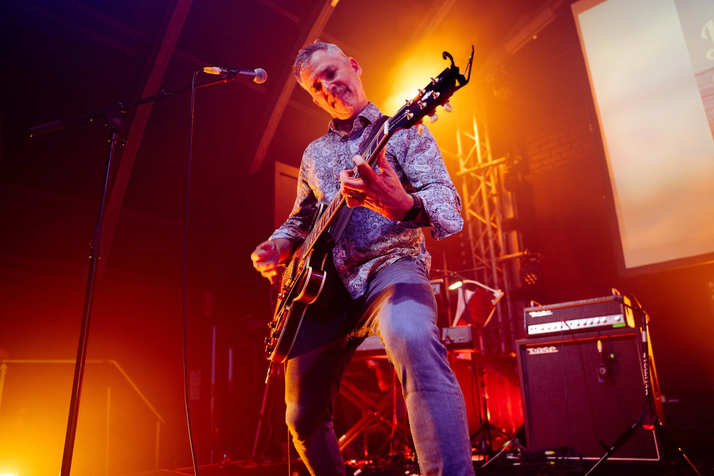 A man playing an electric guitar on stage during a concert with vibrant orange and yellow stage lighting.