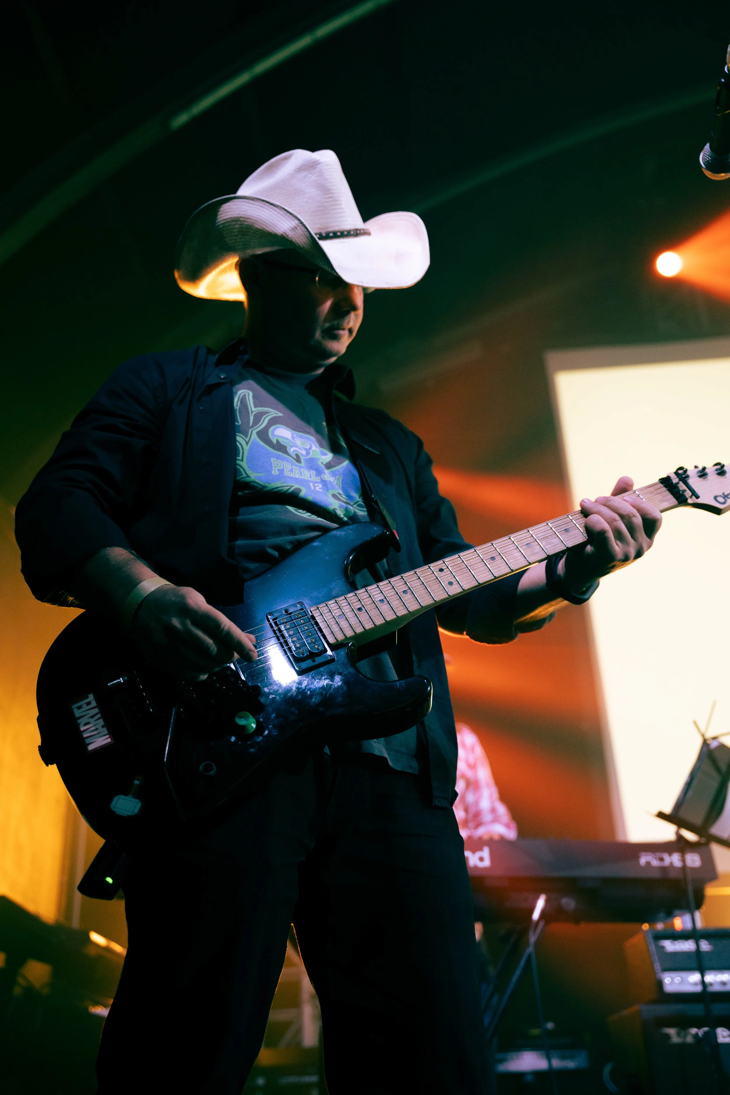 A man wearing a white cowboy hat, glasses, and a black shirt playing an electric guitar on stage with a keyboard in the background and colorful stage lights.