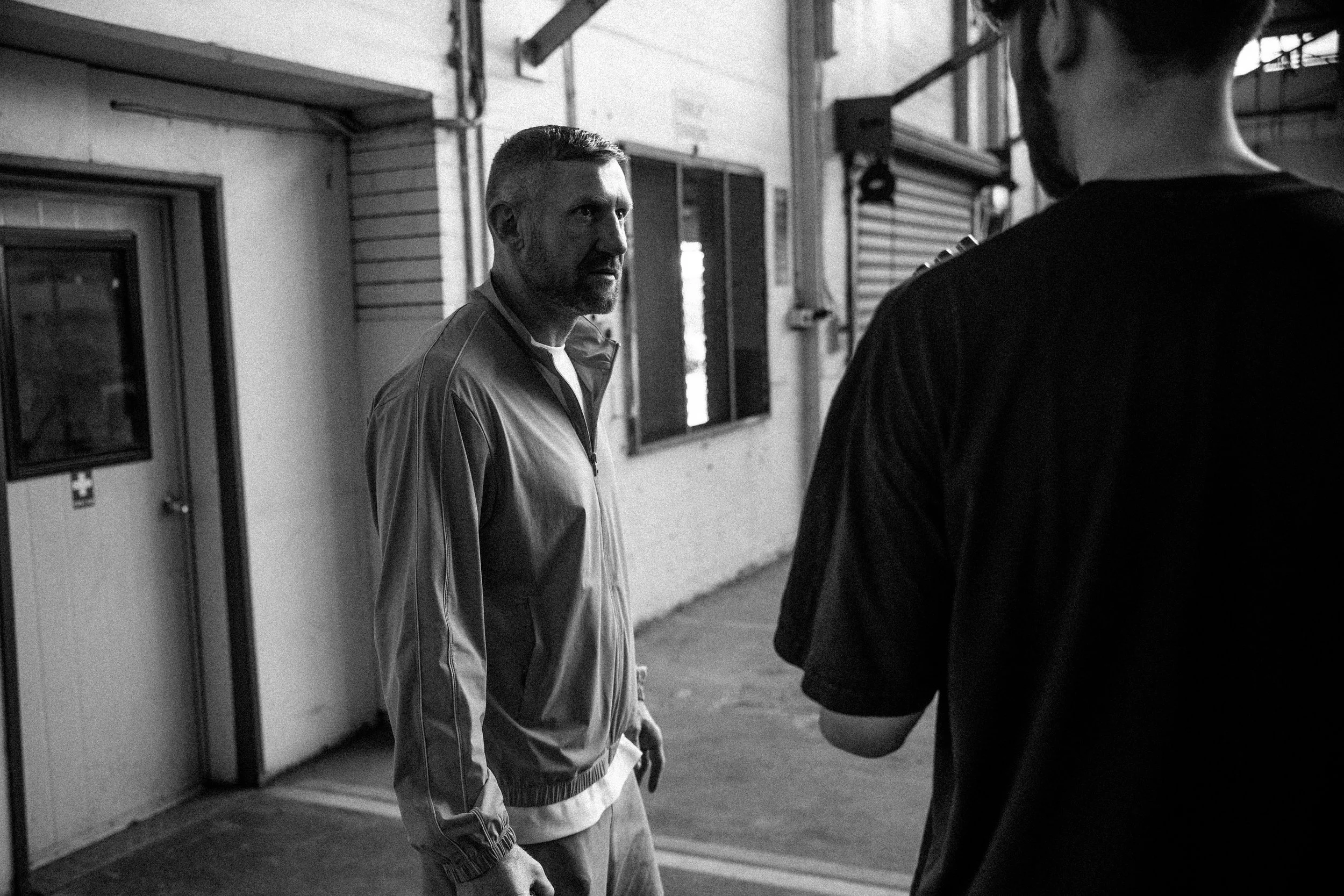 Black and white photo of two men in conversation inside a warehouse or industrial building. One man faces the camera with a serious expression, the other has his back to the camera.