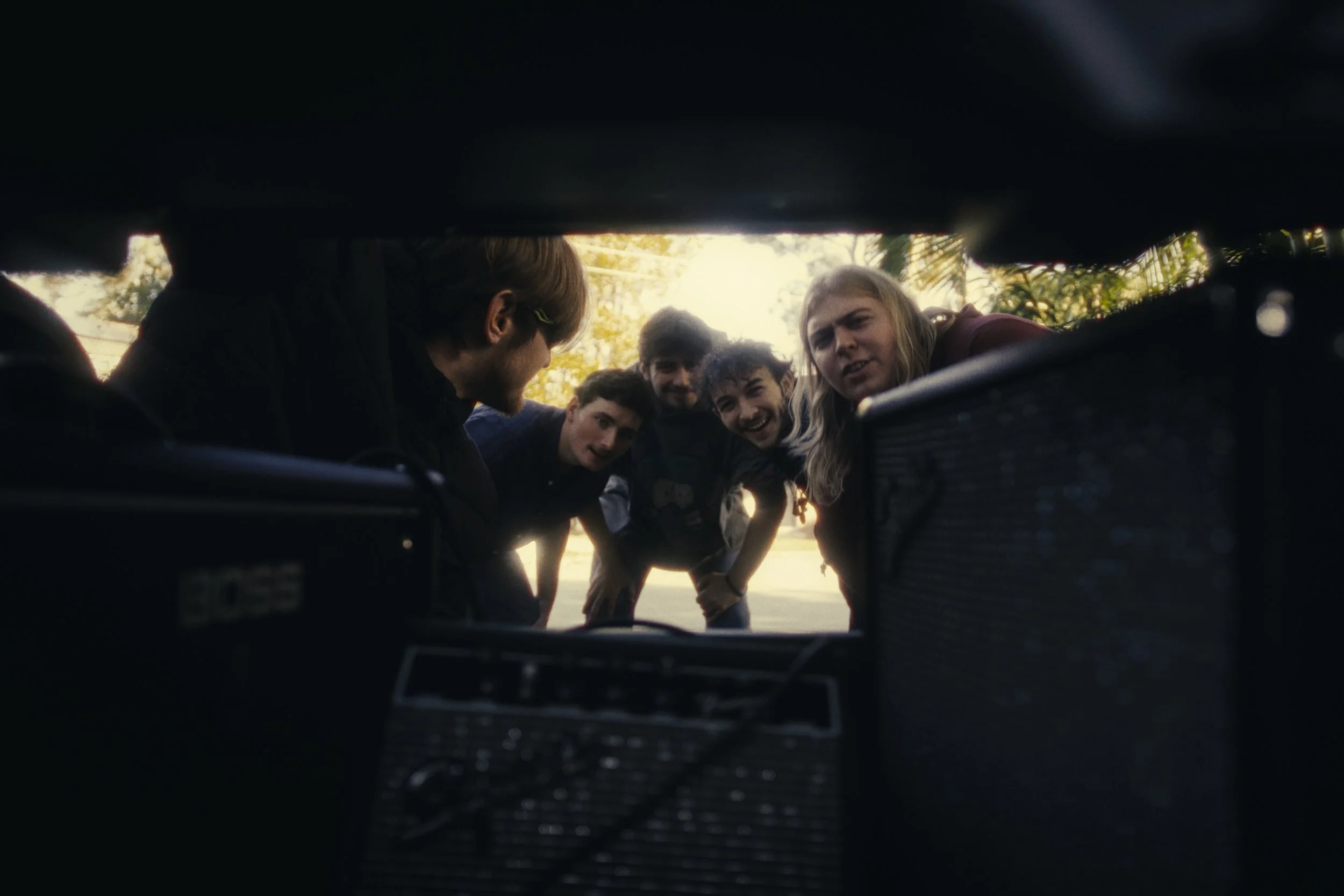 Group of five young people looking into the camera, seen from underneath a vehicle or piece of equipment, with sunlight and trees in the background.