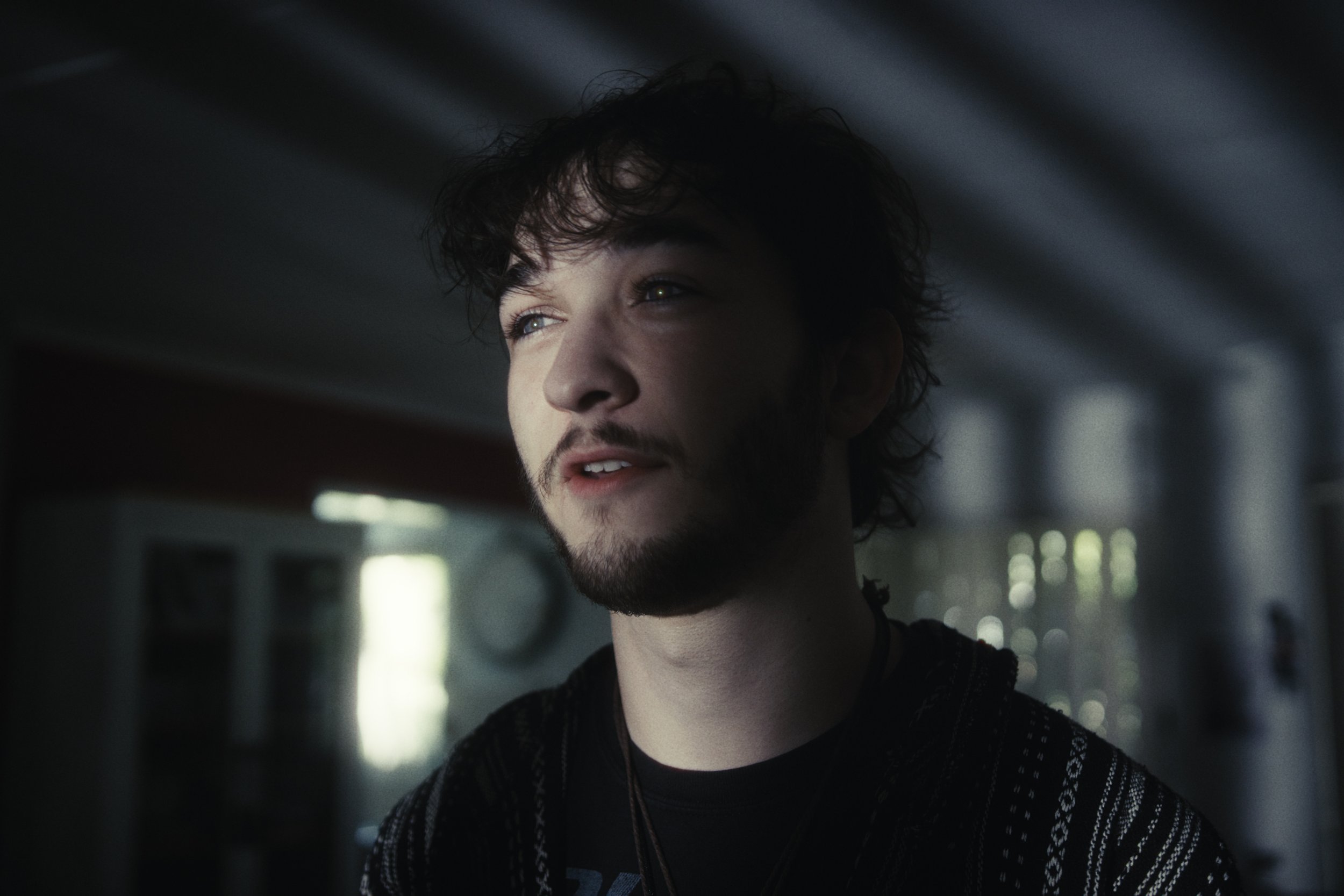 A young man with dark, curly hair and a beard looks off into the distance with a thoughtful expression, indoors with soft natural light coming through windows behind him.