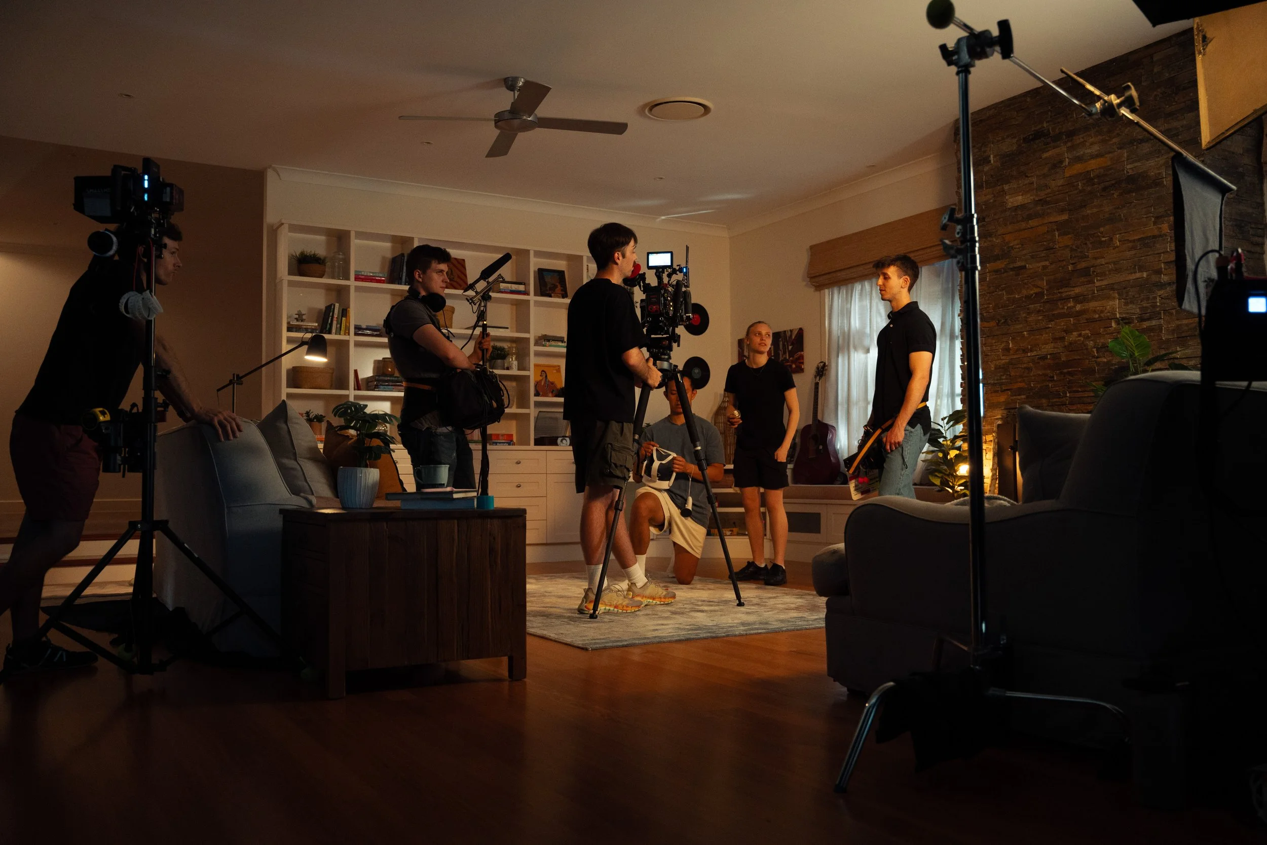 A film crew is filming a scene inside a living room with a brick accent wall, guitar, bookshelf, and plants. The crew includes a camera operator, an assistant, and actors, with equipment and lighting set up around the room.