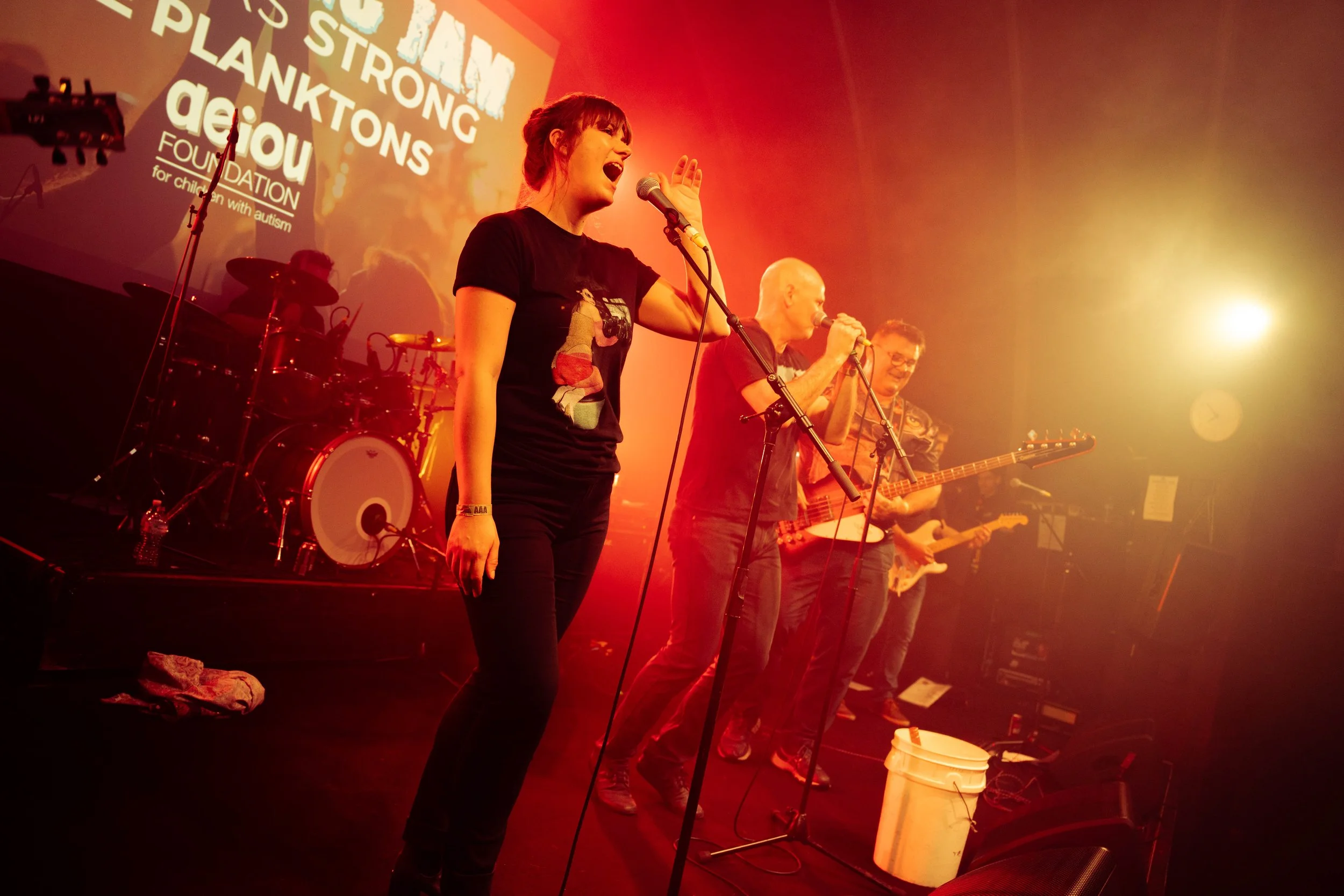 A band performing on stage with orange and yellow lighting, featuring a female singer in the foreground, drums in the background, and guitarists behind her, with a large screen displaying text related to an autism foundation.
