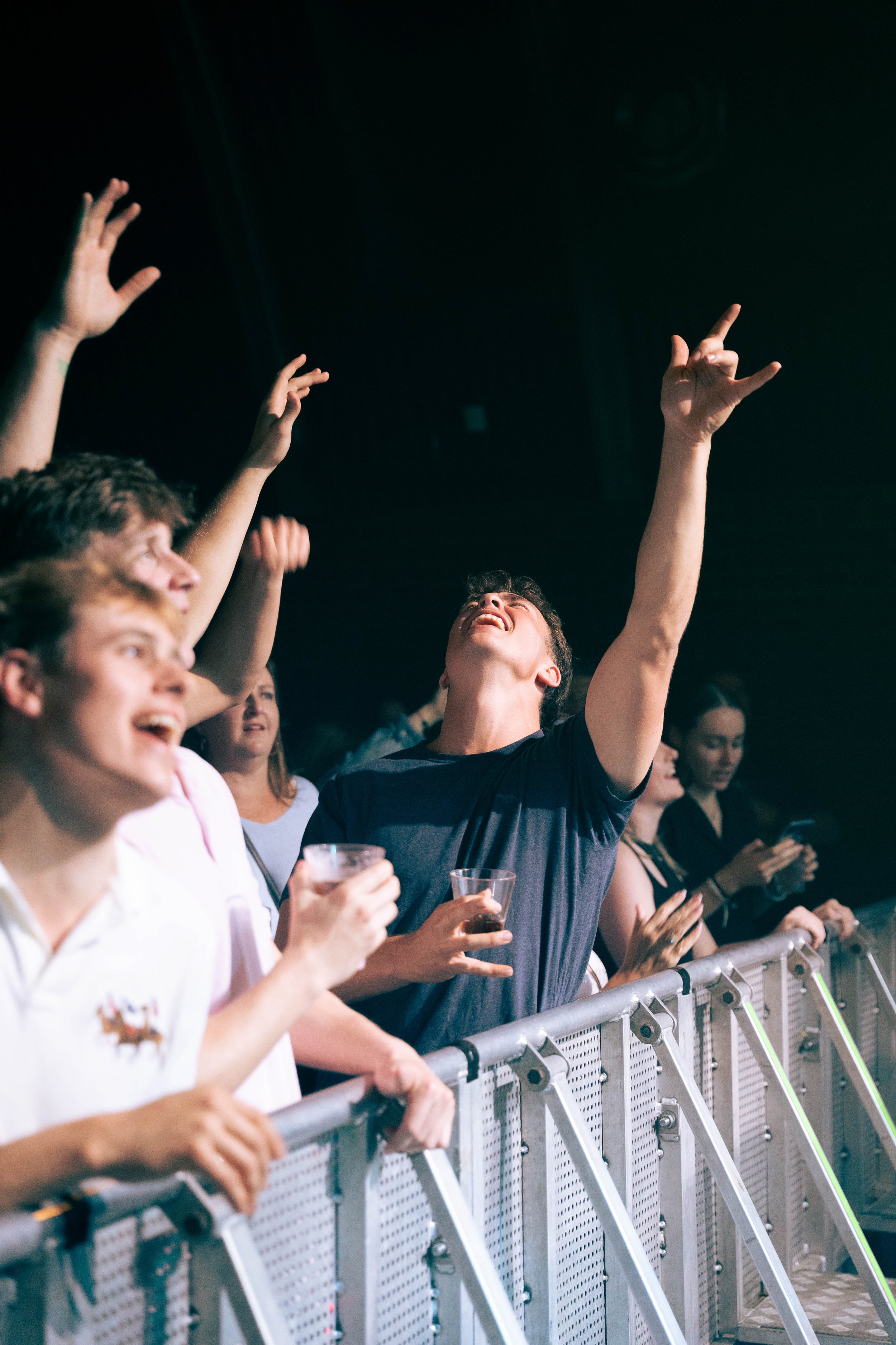Group of young people at a concert or event, with some raising their hands and enjoying the moment, standing behind a barrier.