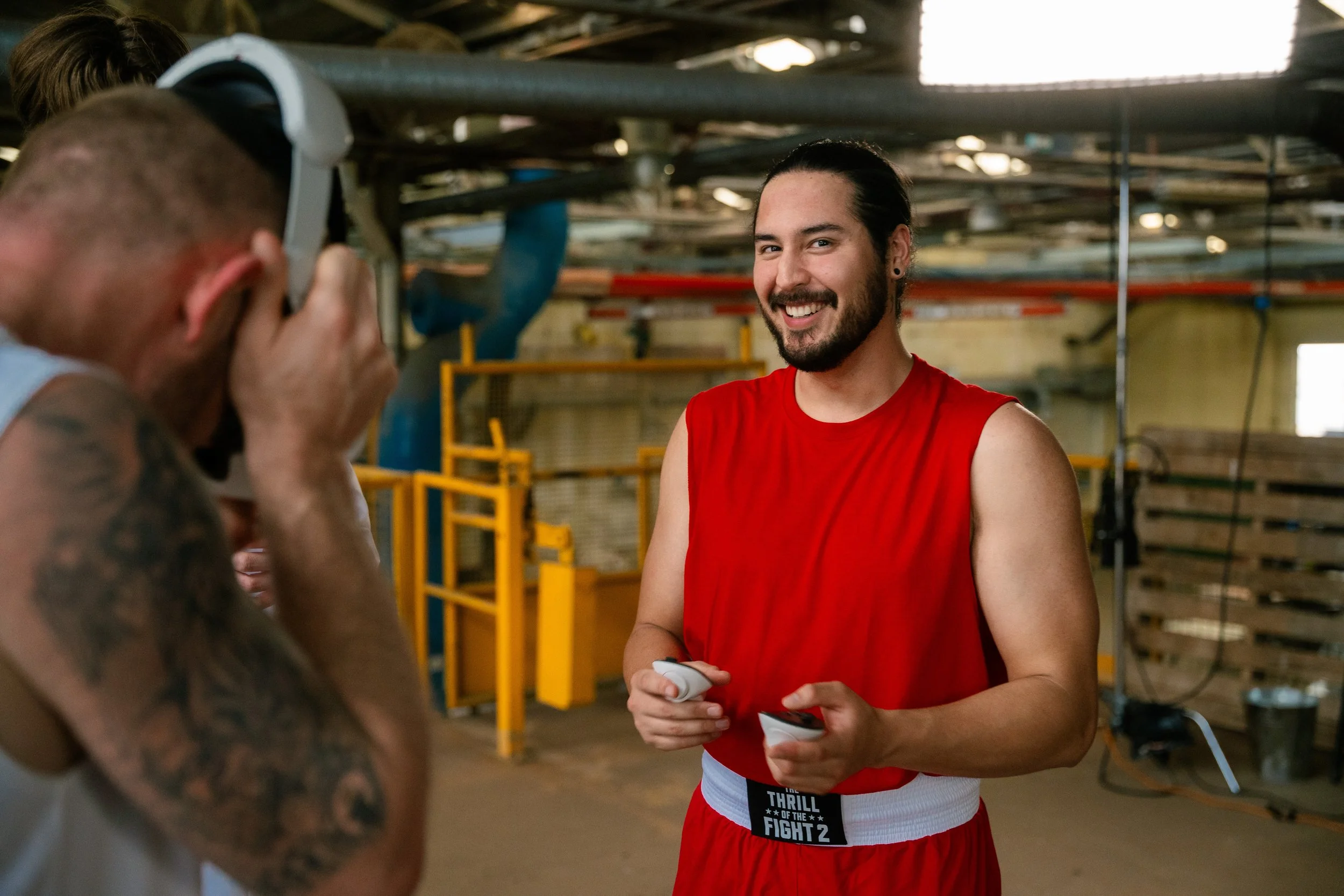 A boxer dressed in red boxing shorts with a white waistband stands smiling while holding a pair of boxing gloves, in a boxing gym with pipes and equipment in the background.