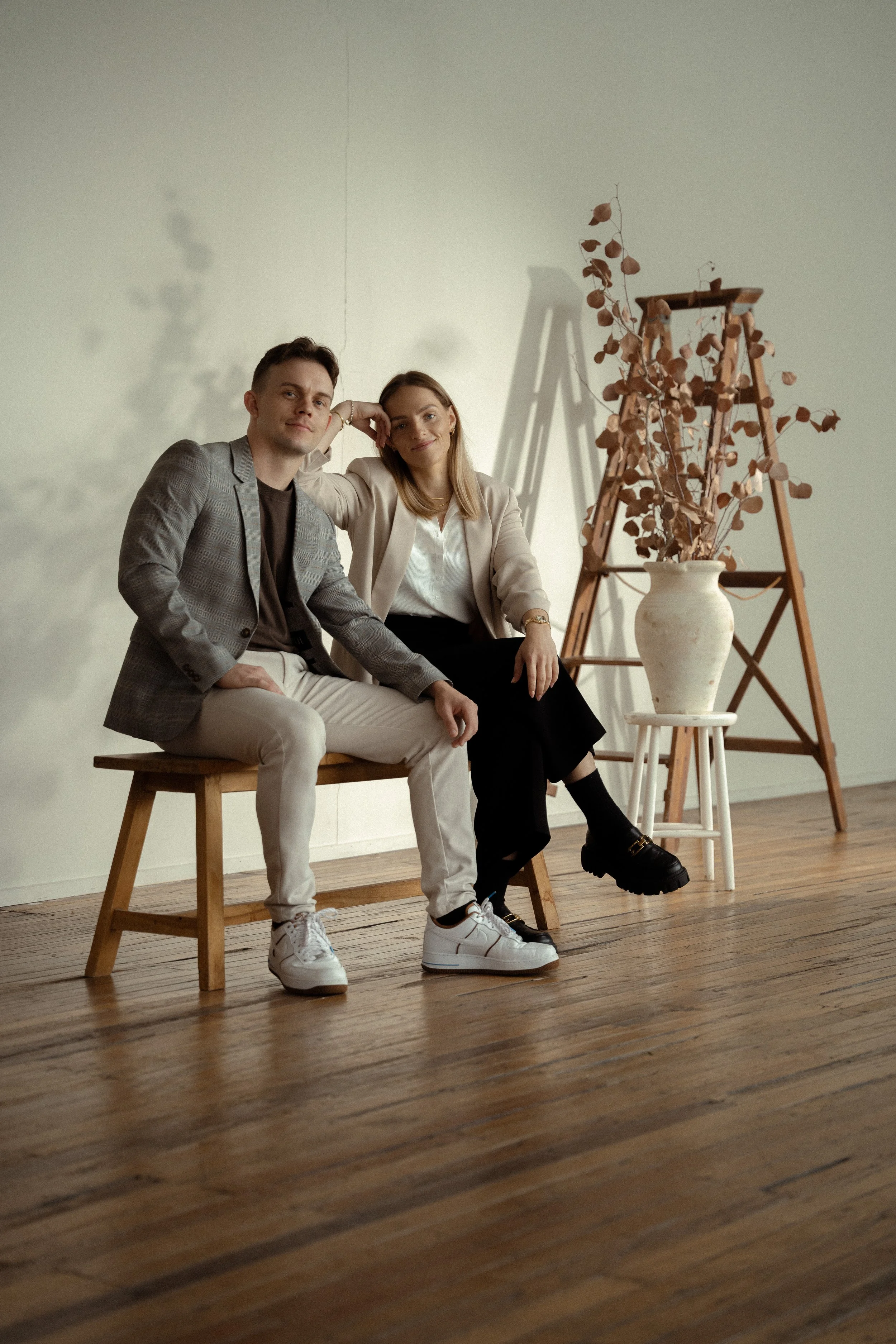 A man and a woman sit on a wooden bench in a softly lit room. The man is dressed in a gray plaid blazer, black shirt, and white pants, while the woman wears a beige blazer, white blouse, and black pants. They sit close together with relaxed expressio