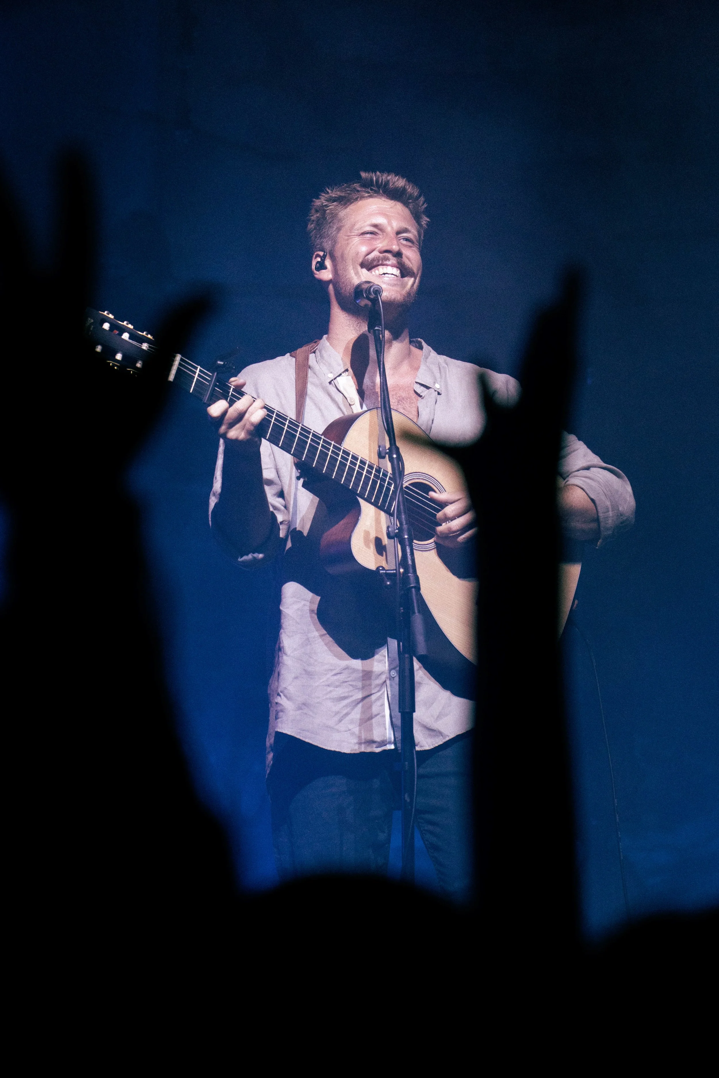 Male musician performing on stage with an acoustic guitar, smiling, with audience hand gestures visible in the foreground.