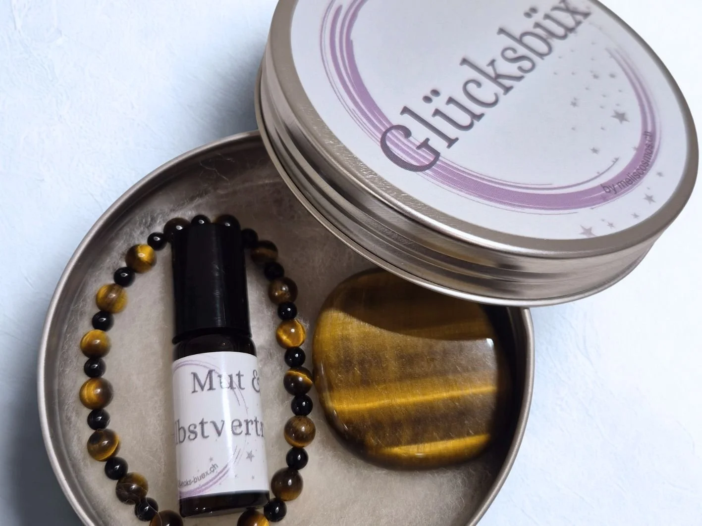 Eine geöffnete Stahlblech-Büchse, die sogenannte Glücksbüx, eine individuelle Geschenkidee. Sie ist gefüllt mit einem Kraftstein Tigerauge, einem Armband aus Tigerauge und Onyx und einem Ölroller (Bergamotte, Zypresse, Rosmarin). Sie kostet CHF 55.-