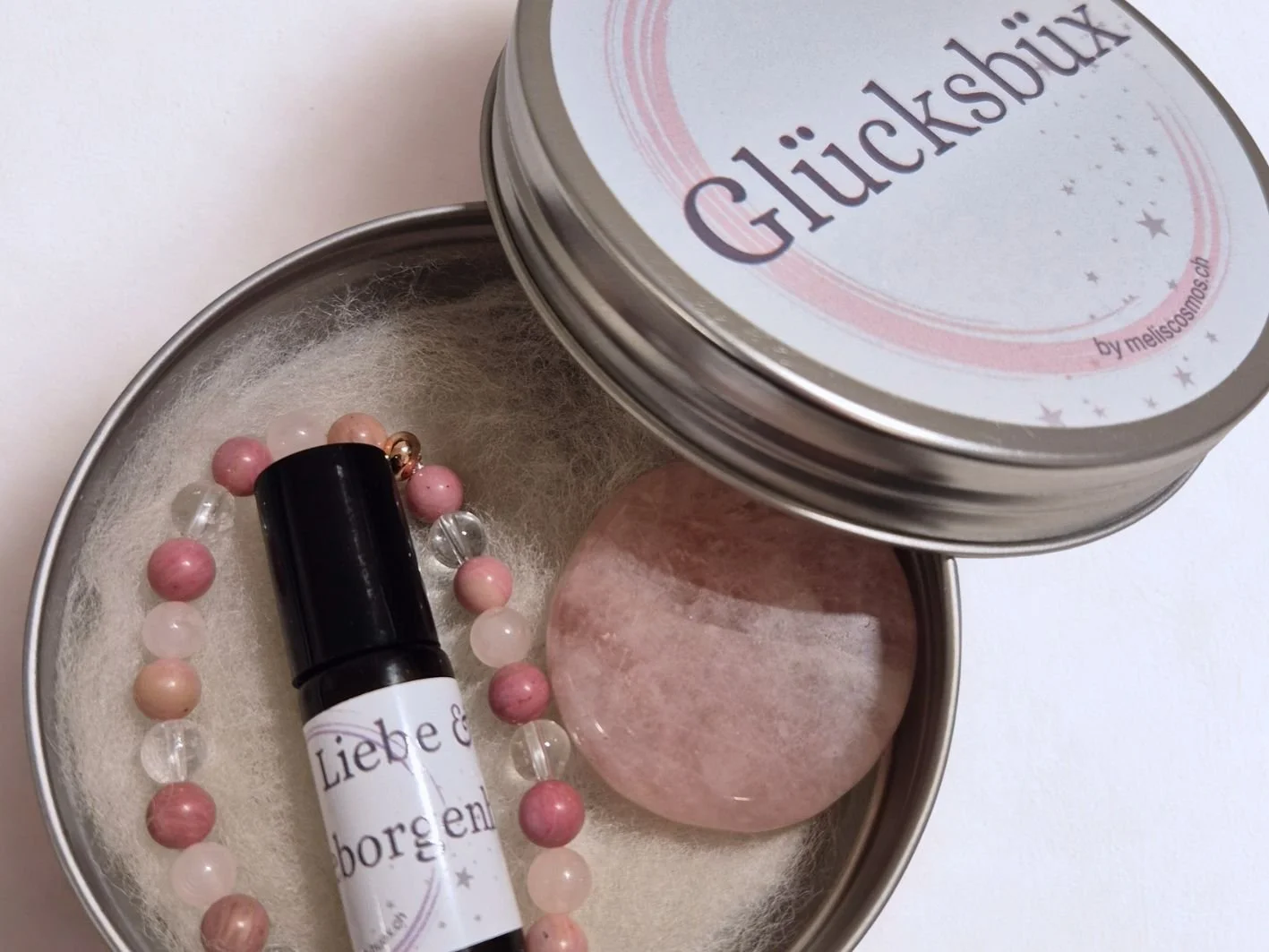 Eine geöffnete Stahlblech-Büchse, die sogenannte Glücksbüx, eine individuelle Geschenkidee. Sie ist gefüllt mit einem Kraftstein Rosenquarz, einem Armband aus Rosenquarz, Rhodonit und Bergkristall und einem Ölroller. Sie kostet CHF 55.-