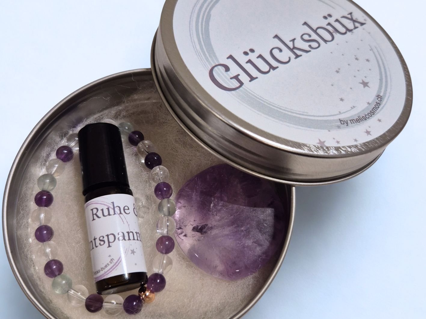 Eine geöffnete Stahlblech-Büchse, die sogenannte Glücksbüx, eine individuelle Geschenkidee. Sie ist gefüllt mit einem Kraftstein Amethyst, einem Armband (Bergkristall, Amethyst, Fluorit) und einem Ölroller. Sie kostet CHF 55.-