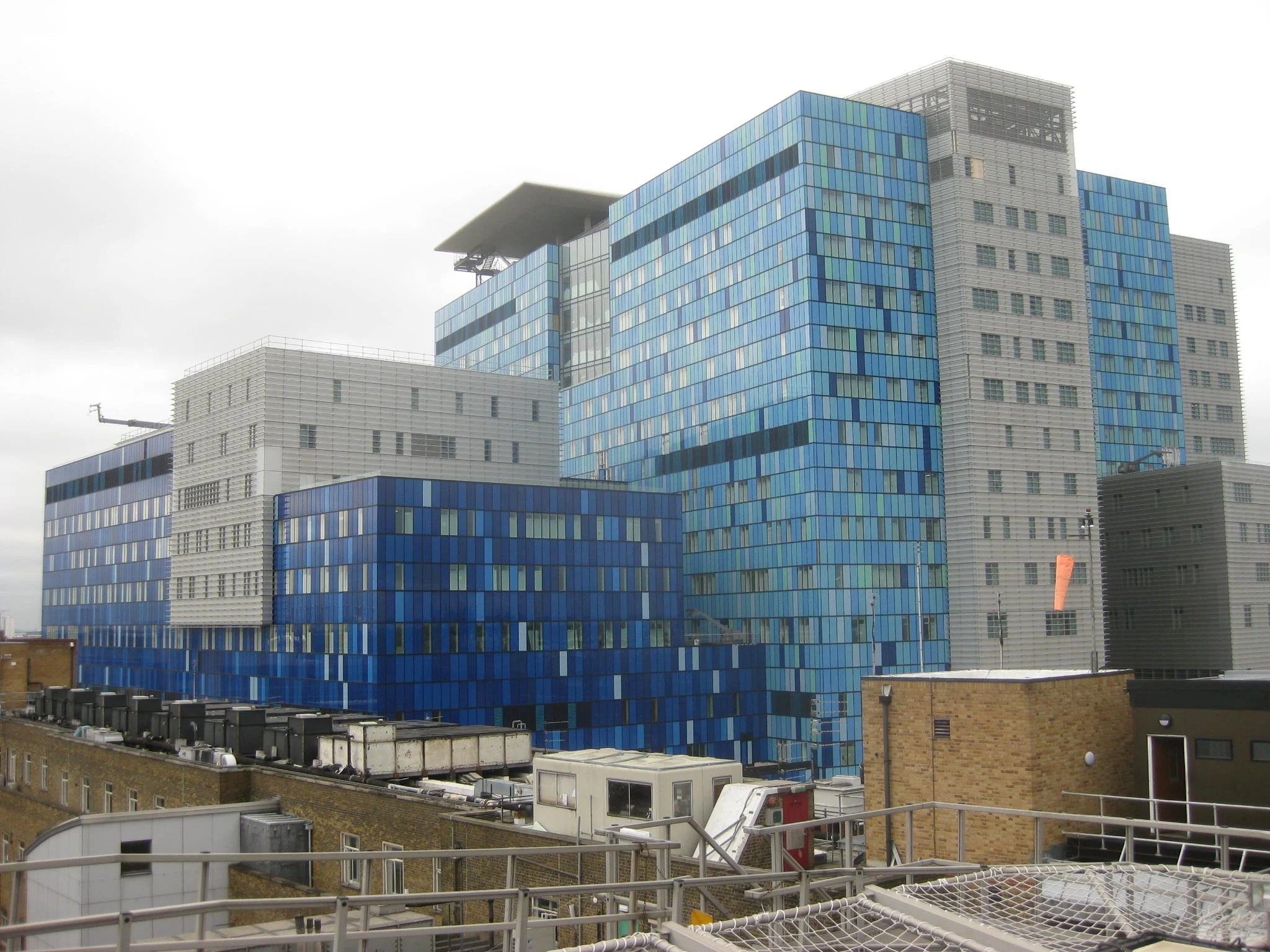 Modern skyscraper with blue glass windows and multiple levels, featuring architectural details and surrounding rooftops under a cloudy sky.