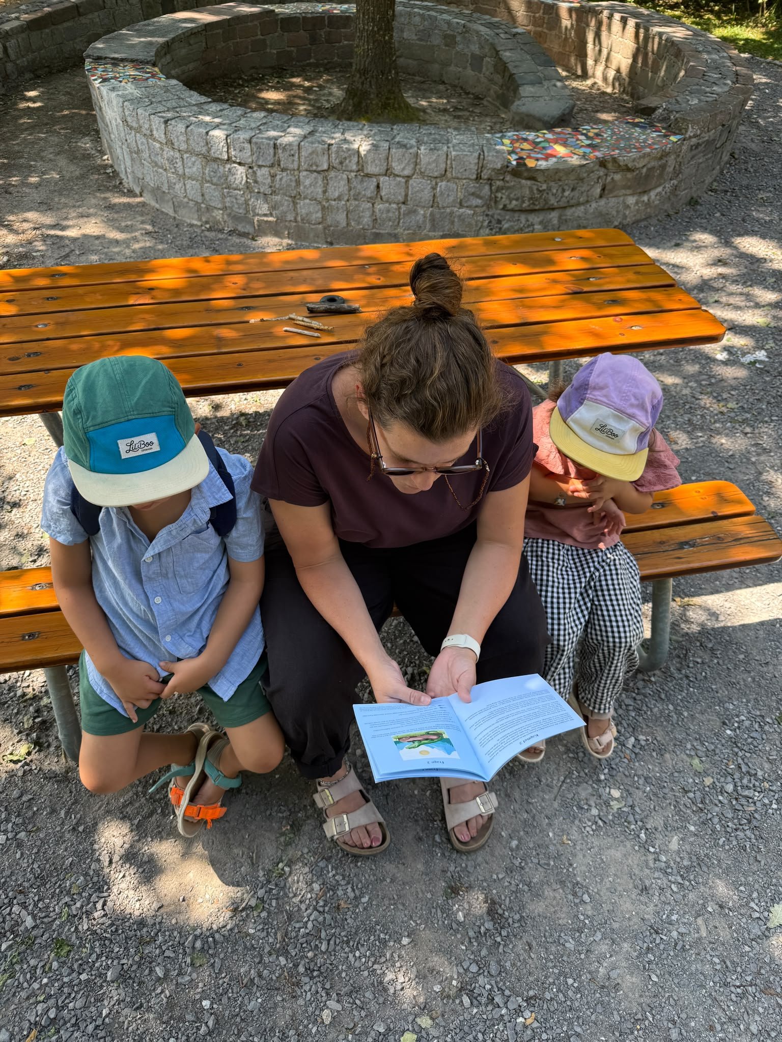 Eine Frau sitzt mit zwei Kindern in der Natur an einem Tisch und liest ihnen ein Kapitel aus einem Family Adventure to go vor.