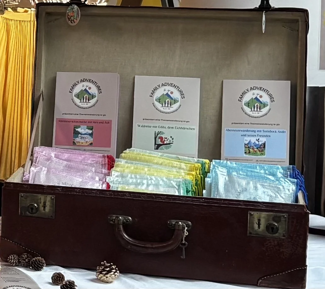 Ein offener Koffer auf einem Tisch, gefüllt mit bunten Stofftaschen und Informationsblättern von 'Family Adventures', die verschiedene Themen und Aktivitäten für Kinder und Familien präsentieren.