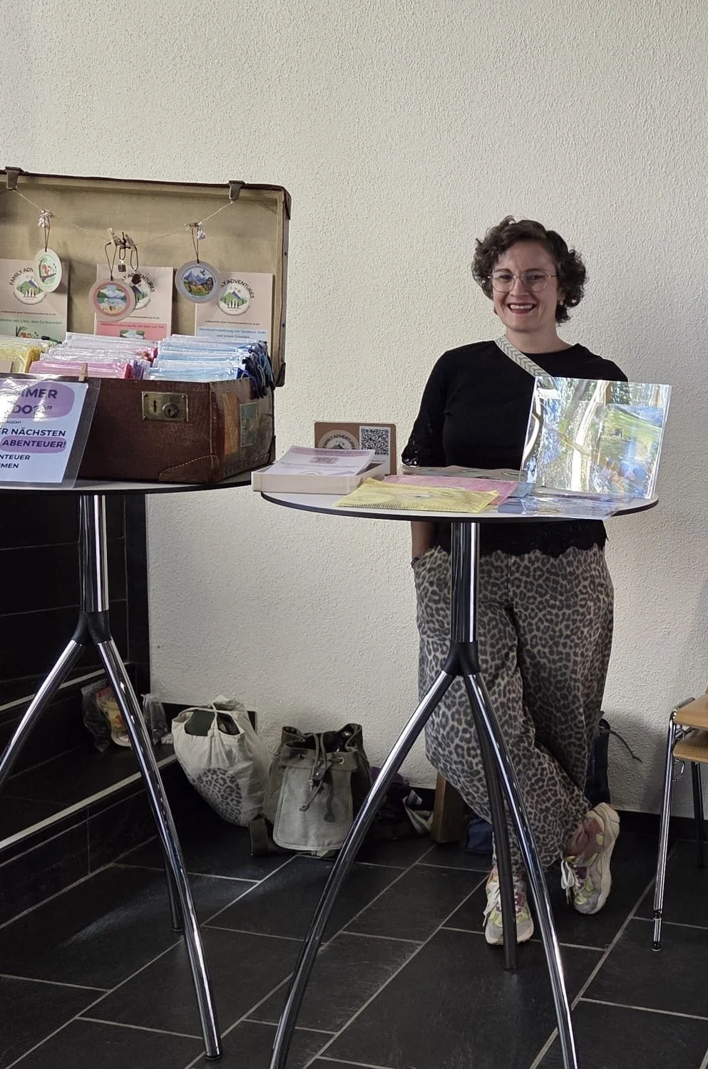 Die Inhaberin von Family Adventures to go steht hinter einem runden Stehtisch mit Flyern und Broschüren, im Hintergrund ein offener Koffer mit den Adventures