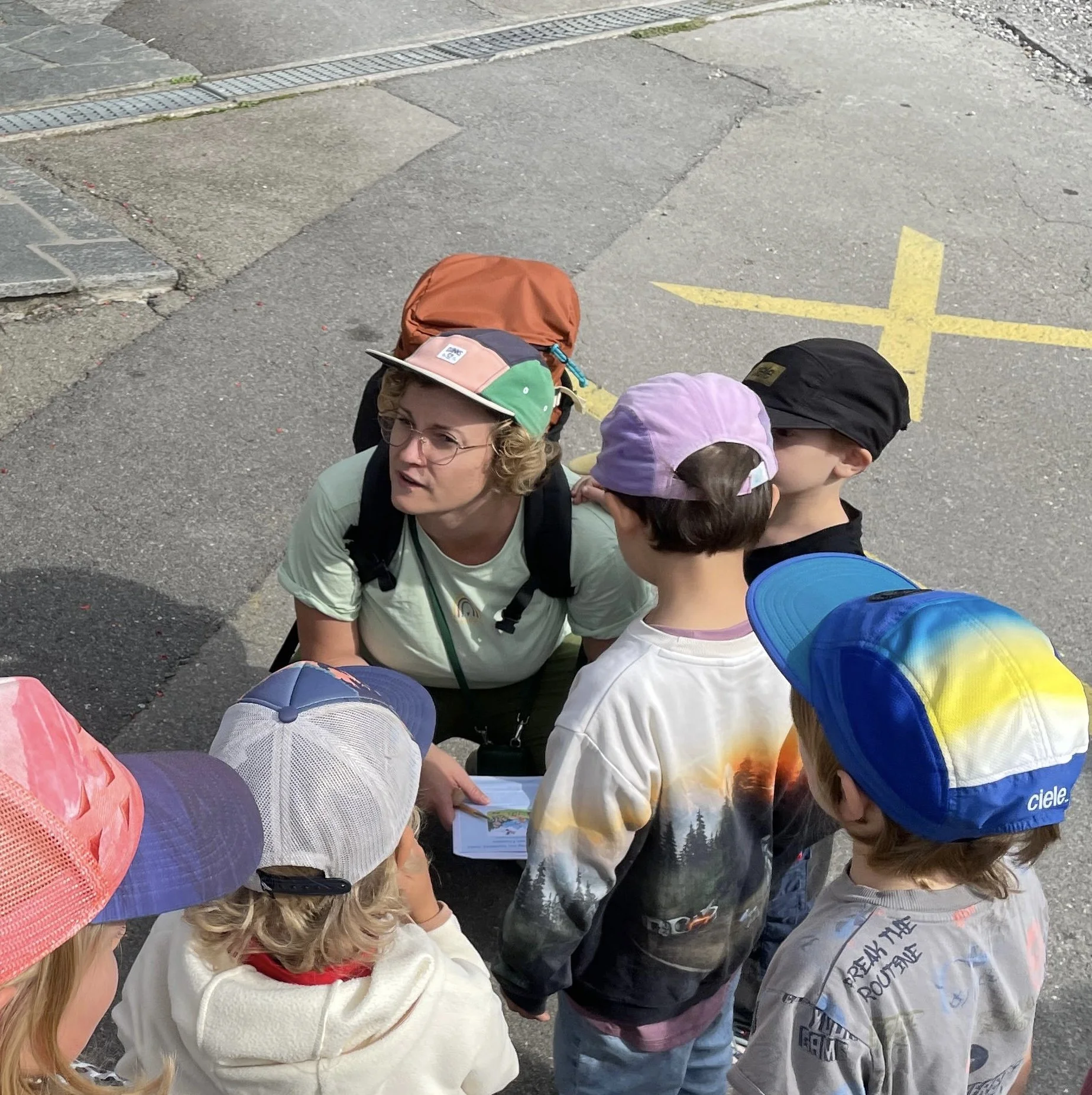 Eine Gruppe Kinder und eine Erwachsene Person stehen auf einem Bürgersteig. Die Erwachsene Personzeigt auf eine Karte oder ein Blatt und erklärt den Kindern etwas. Alle tragen Hüte und Rucksäcke, vermutlich beim Ausflug oder bei einer Schulaktivität.