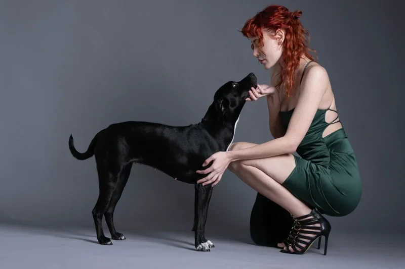 A woman with red hair in a green dress crouches down to pet a black dog against a gray background.