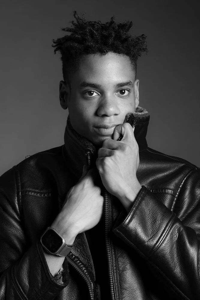 A young man with styled hair, wearing a leather jacket and a smartwatch, is holding his collar with one hand and touching his face with the other, looking directly at the camera in a black and white portrait.