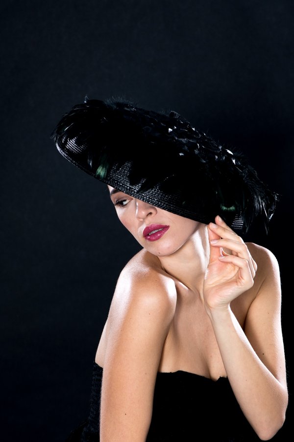 A woman with a black feathered hat covering her eyes, posing against a dark background.