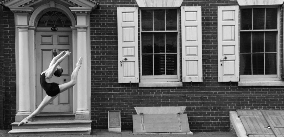 Ballet dancer performing a leap in front of a brick building with a door and a window with shutters.
