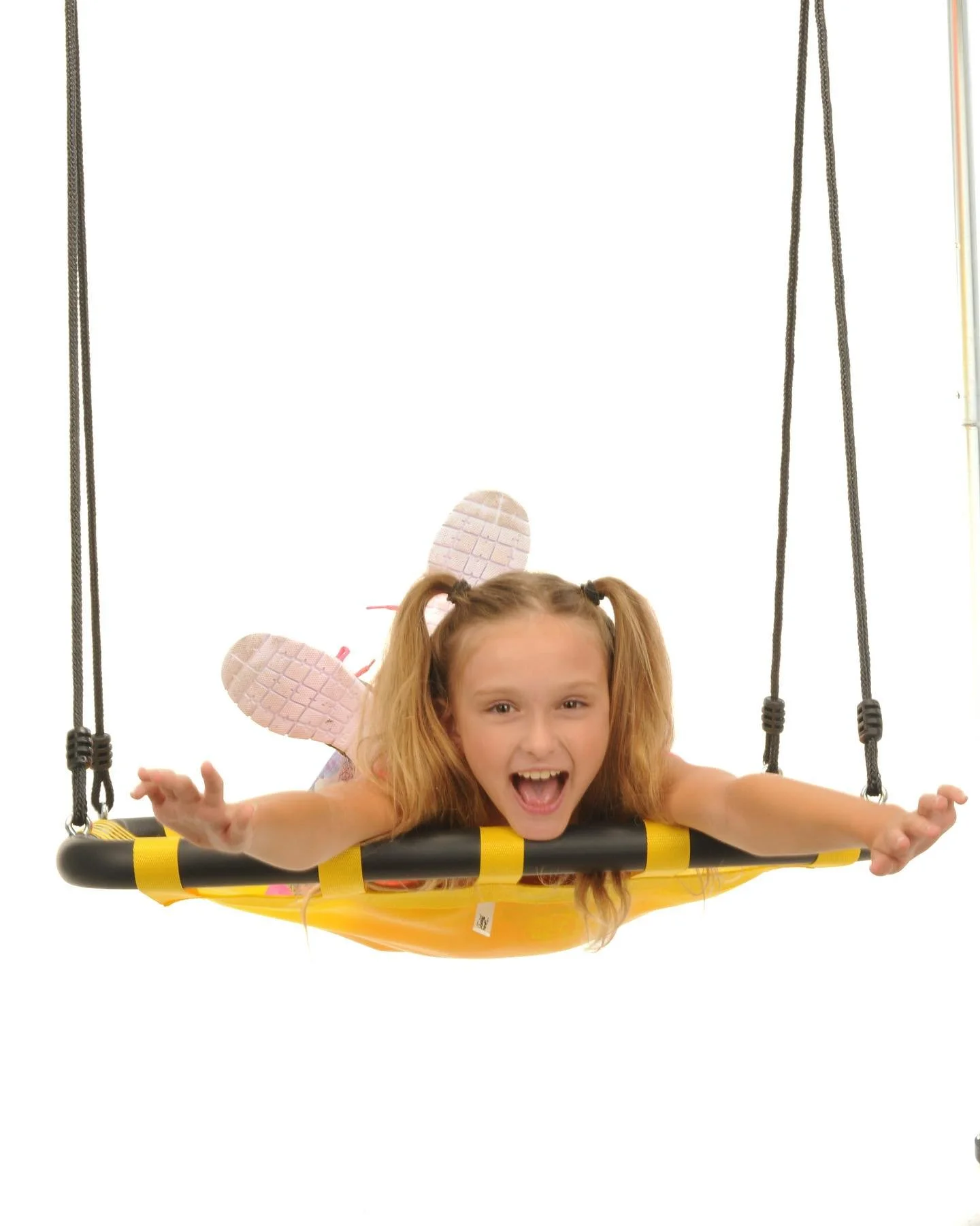 A young girl with pigtails and fairy wings lying on a swing with a yellow seat, smiling and reaching out with both arms, against a white background.