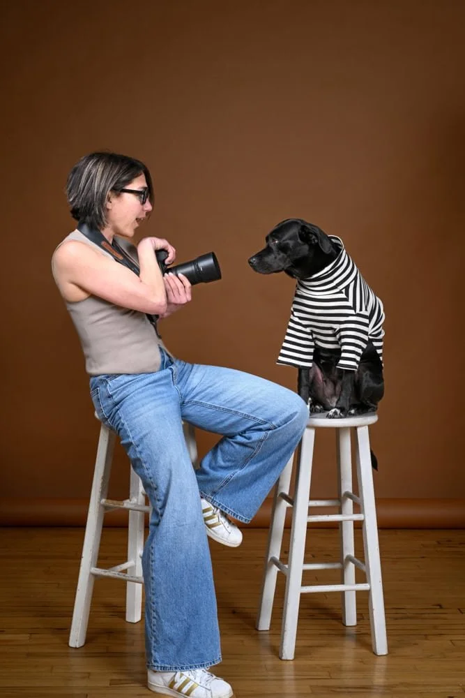 A woman with glasses taking a photo of a dog dressed in a black and white striped shirt sitting on a stool in a photography studio with a brown backdrop.