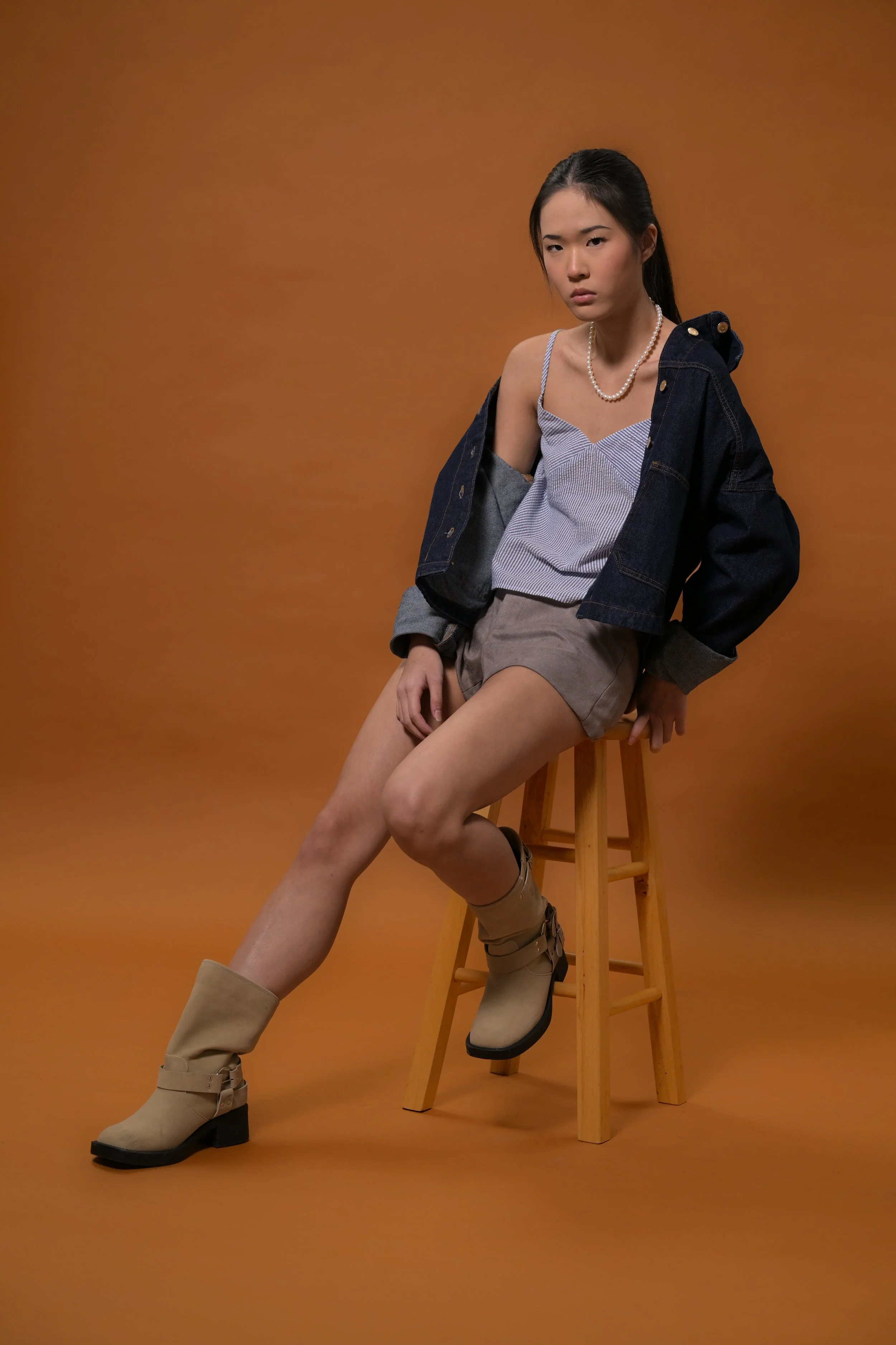 Young woman sitting on a wooden stool against a brown background, wearing a denim jacket, striped top, tan shorts, and beige boots.