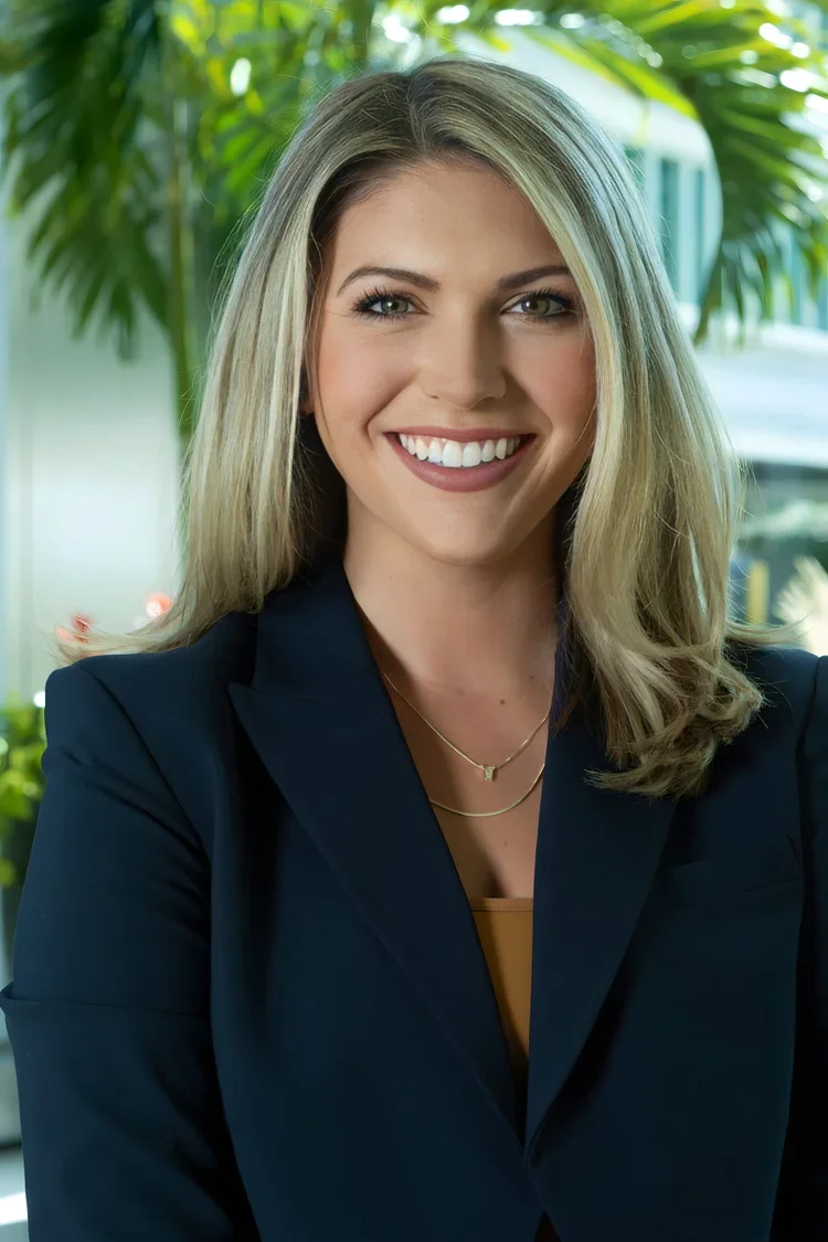 Smiling woman with blonde hair, wearing a navy blazer and gold layered necklaces, standing in front of green plants.