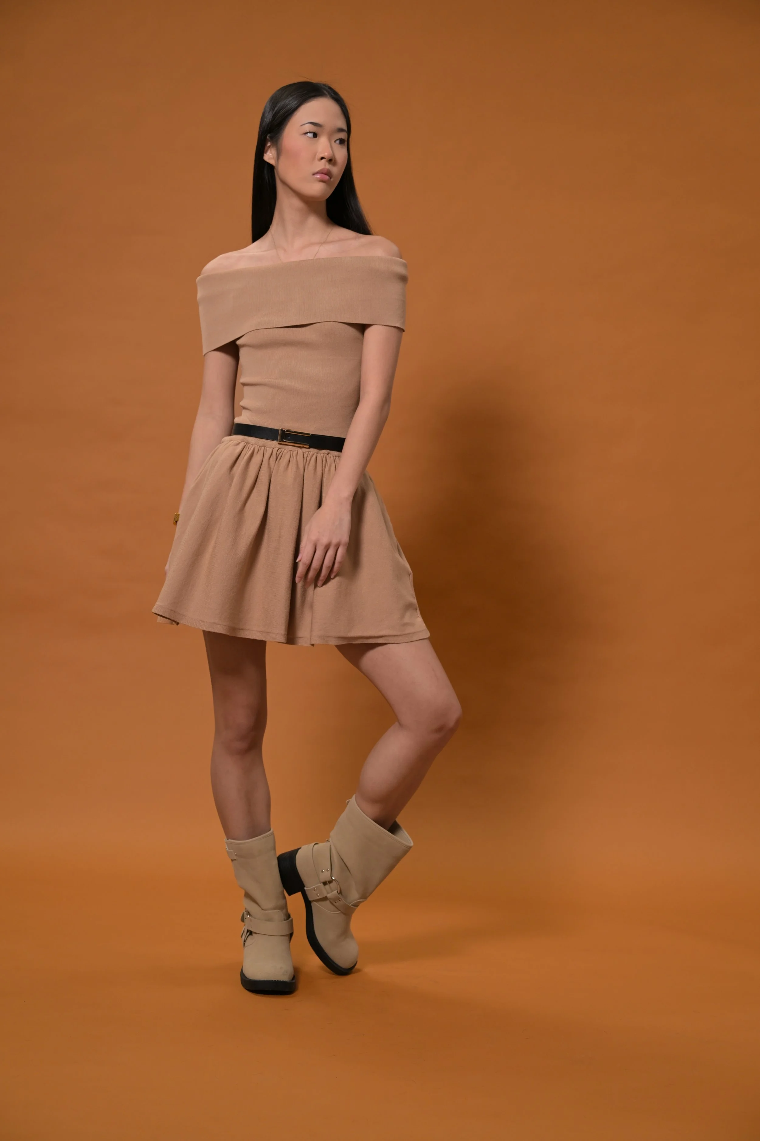 A woman with long black hair dressed in a beige off-the-shoulder top, beige skirt, and beige boots, stands against a brown background.